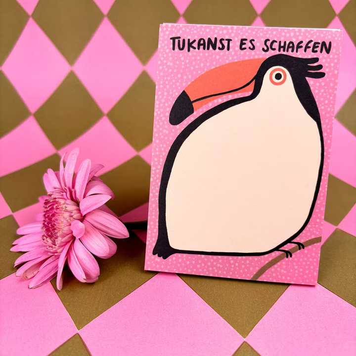 Eine Grußkarte mit einem Cartoon-Tukan und dem deutschen Text TUKANST ES SCHAFFEN steht neben einem rosa Gänseblümchen auf einem rosa-gold karierten Hintergrund.