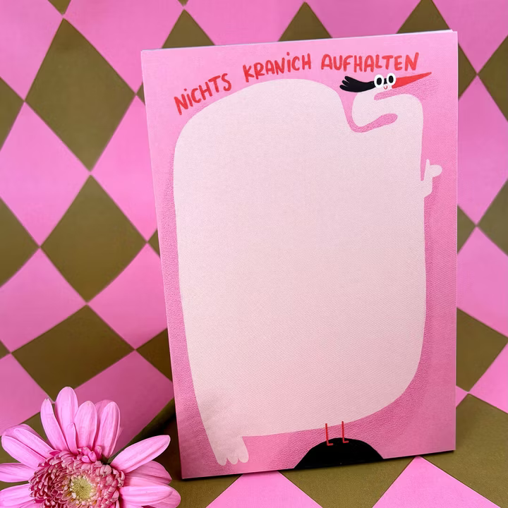 Ein rosafarbener Notizblock mit einem Cartoon-Storch und dem deutschen Text NICHTS KRANICH AUFHALTEN am oberen Rand, platziert auf einer rosa und oliv karierten Oberfläche mit einer rosa Blume in der Nähe.