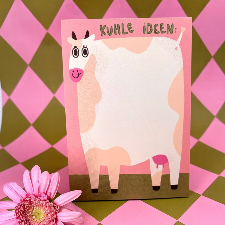 Ein rosa-weißer Notizblock mit einem Cartoon-Kuh-Motiv und dem Titel Kuhle Ideen. Der Hintergrund besteht aus rosa und braunen Rauten, und in der Ecke sitzt eine rosa Blume.