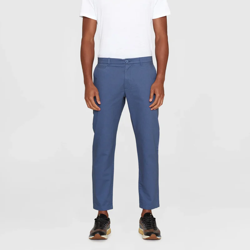 Eine Person, die ein einfaches weißes T-Shirt, eine blaue Slim-Fit-Chino und schwarze Turnschuhe mit weißen Sohlen trägt und vor einem einfachen weißen Hintergrund steht.