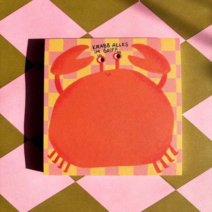 Eine rosafarbene und orangefarbene Zeichnung einer lächelnden Krabbe mit erhobenen Krallen befindet sich auf einer karierten quadratischen Karte. Der deutsche Text darüber lautet KRABB ALLES IM GRIFF. Die Karte ruht auf einer rosa und grün karierten Fläche.