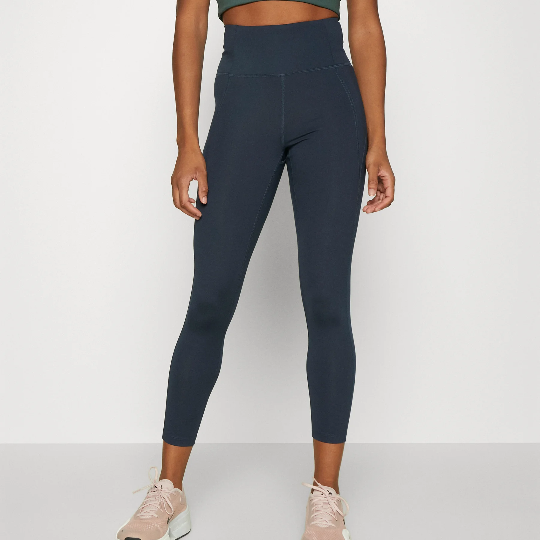 Eine Person, die hochtaillierte, dunkelblaue Leggings, ein dunkelgrünes Crop-Top und hellrosa Sportschuhe trägt, steht vor einem schlichten weißen Hintergrund. Ihr Kopf ist auf dem Bild nicht zu sehen.