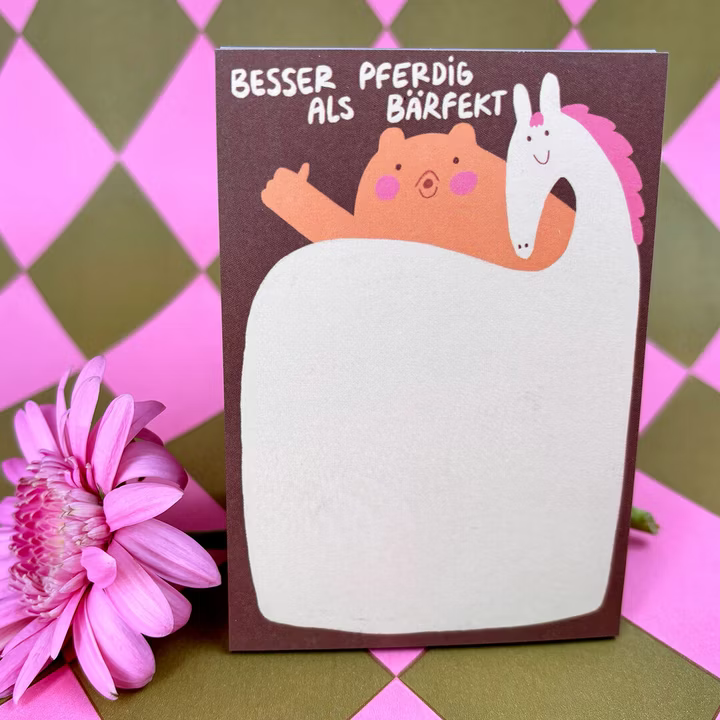 Ein Notizblock mit einem Cartoon-Bären und einem Pferd, auf dem der deutsche Satz Besser pferdig als bärfekt steht. Vorne liegt eine rosafarbene Blume, und der Hintergrund hat ein rosa und olivgrünes Rautenmuster.