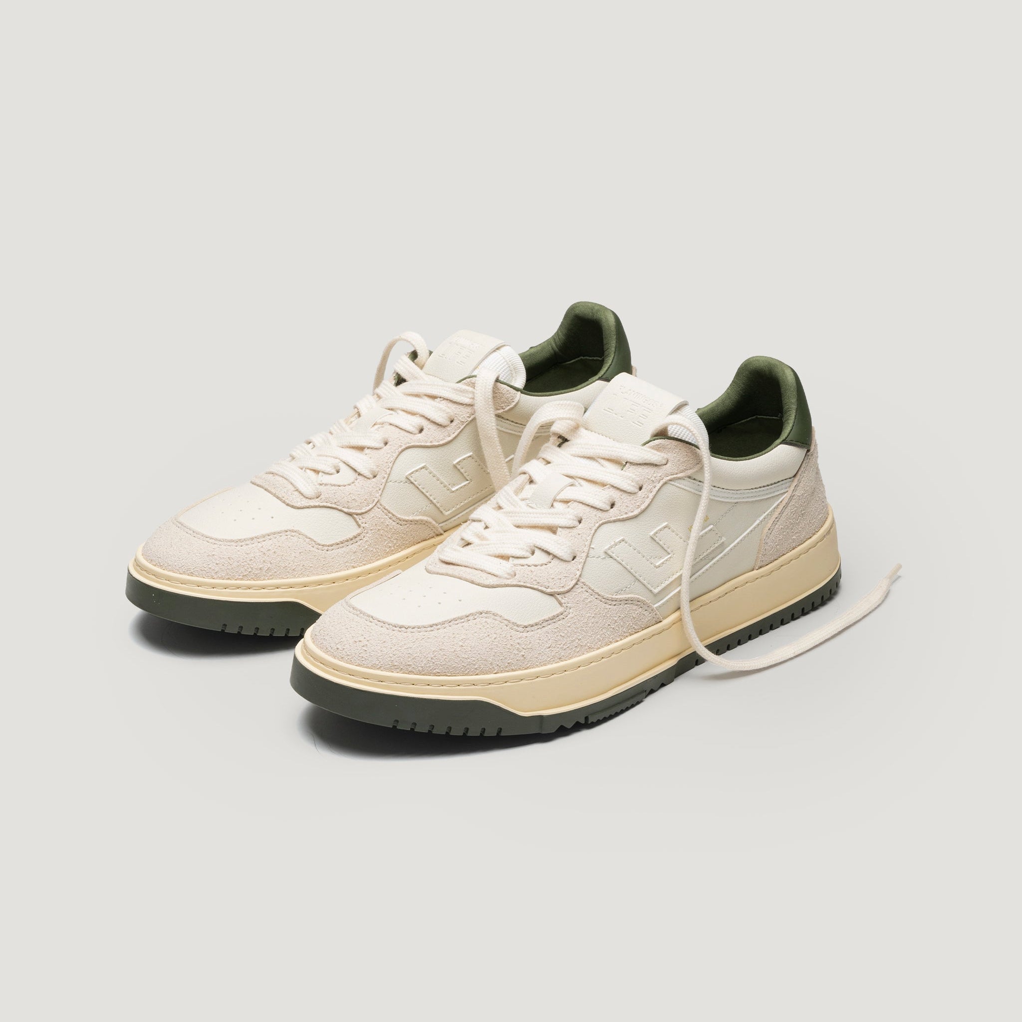 Ein Paar cremefarbene Turnschuhe mit cremefarbener Sohle, dunkelgrünem Innenfutter und schwarzer Laufsohle, dargestellt auf hellem Hintergrund, wobei ein Schnürsenkel offen ist.