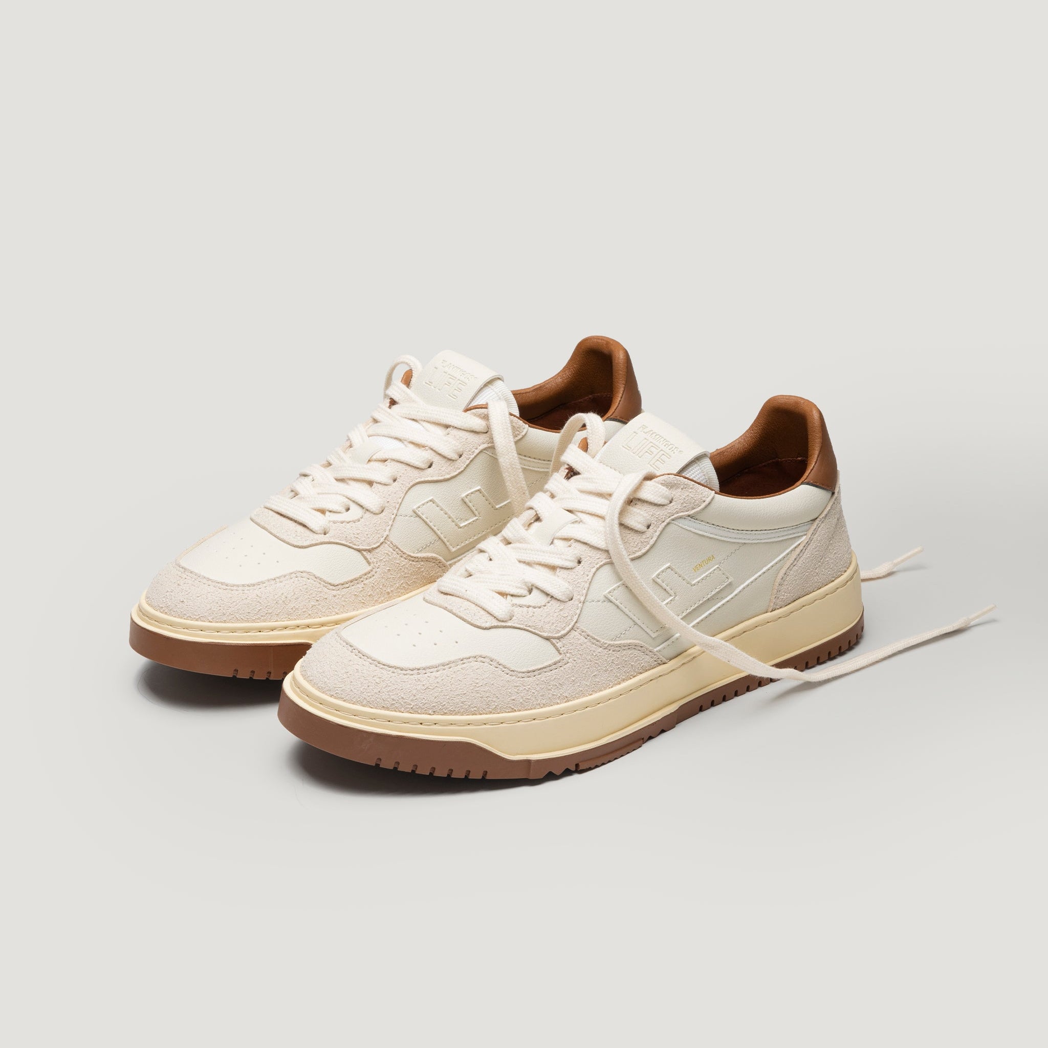 Ein Paar beige- und cremefarbene Low-Top-Sneakers mit braunen Sohlen und Akzenten vor einem schlichten hellen Hintergrund. Ein Schnürsenkel ist gelöst und lose nach vorne drapiert.