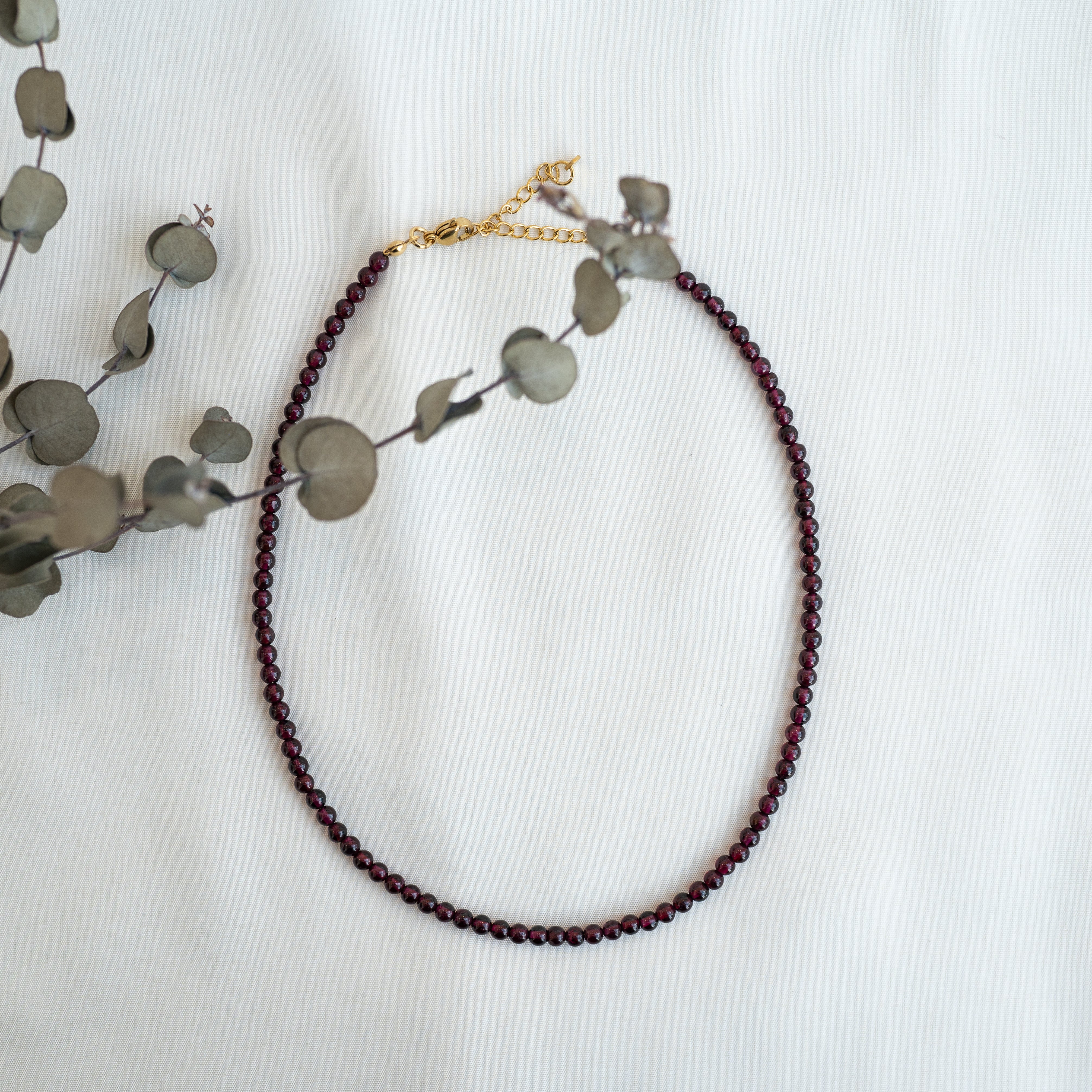 Eine zarte Perlenkette mit dunkelroten Perlen und einem goldenen Verschluss ist auf einer weißen Fläche ausgestellt, die teilweise von einem Zweig getrockneten Eukalyptus überlagert wird.