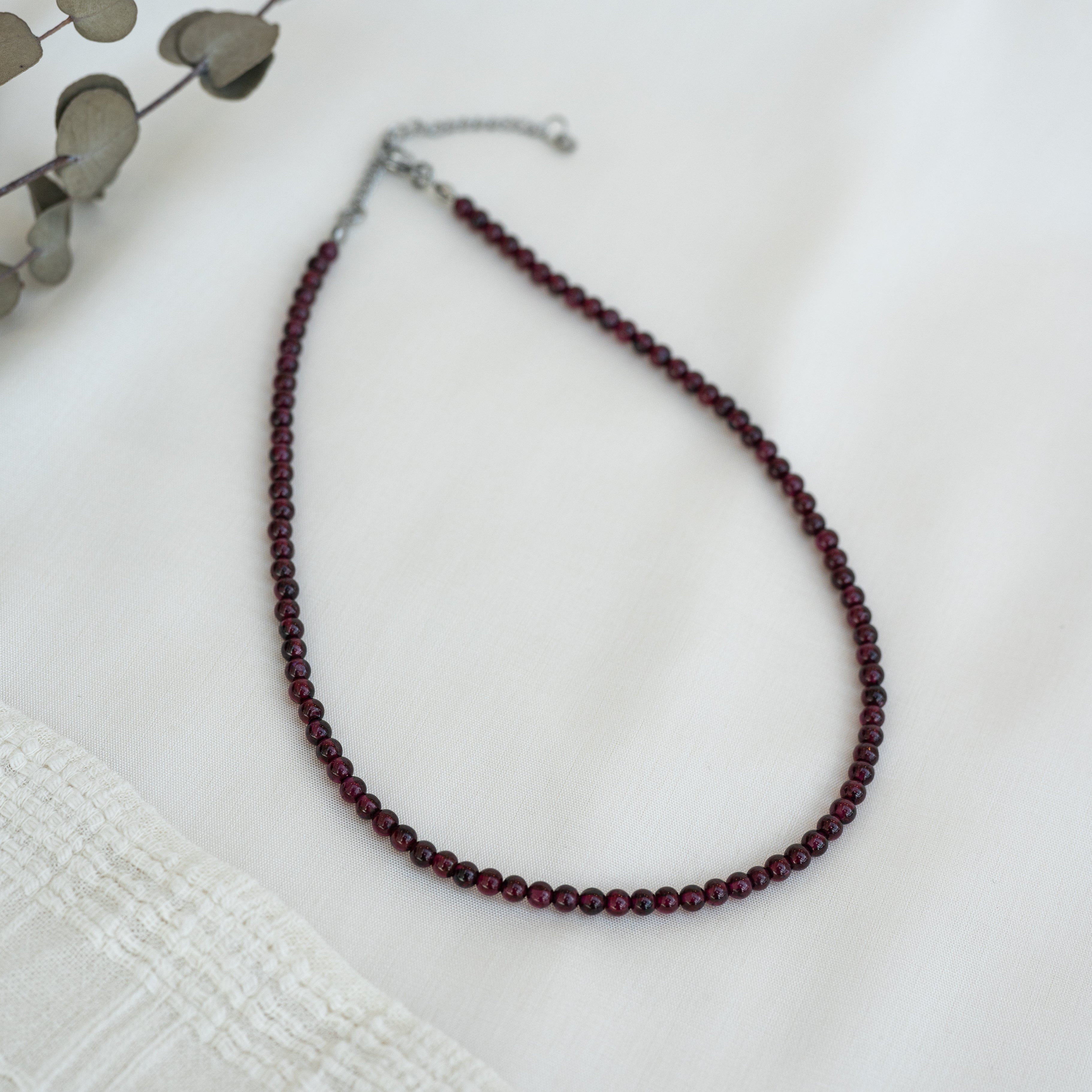 Eine einsträngige Halskette aus kleinen, runden, dunkelroten Perlen liegt auf einer weißen Stofffläche neben getrockneten Eukalyptusblättern und strukturiertem Stoff.