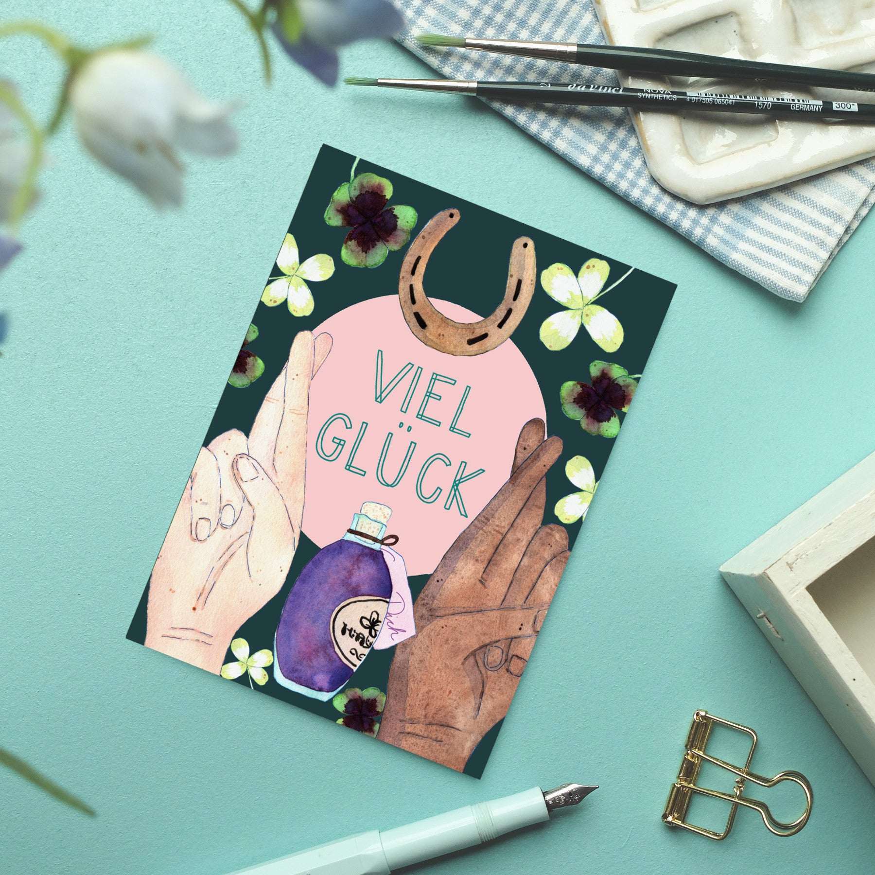 Eine Postkarte auf einer blaugrünen Oberfläche zeigt drei illustrierte Hände, ein Hufeisen, Kleeblätter und eine Zaubertrankflasche. In der Mitte steht "Viel Glück". Kunstzubehör und eine Blume rahmen die Szene ein.