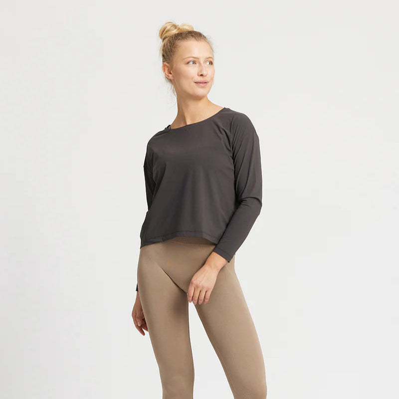 Eine Frau mit blondem Haar in einem Dutt trägt ein langärmeliges schwarzes Oberteil und taupefarbene Leggings. Sie steht vor einem schlichten weißen Hintergrund und blickt mit einem leichten Lächeln zur Seite.
