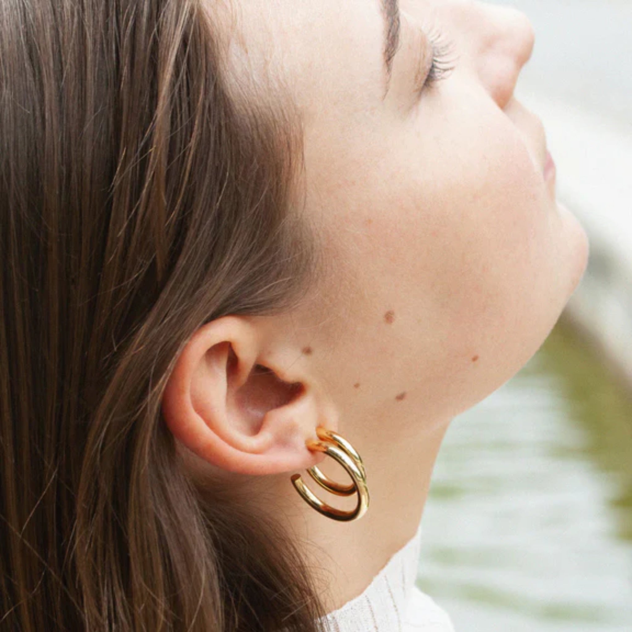 Nahaufnahme eines Ohres mit goldenen Creolen, im Hintergrund ein unscharfer Wasserbereich.
