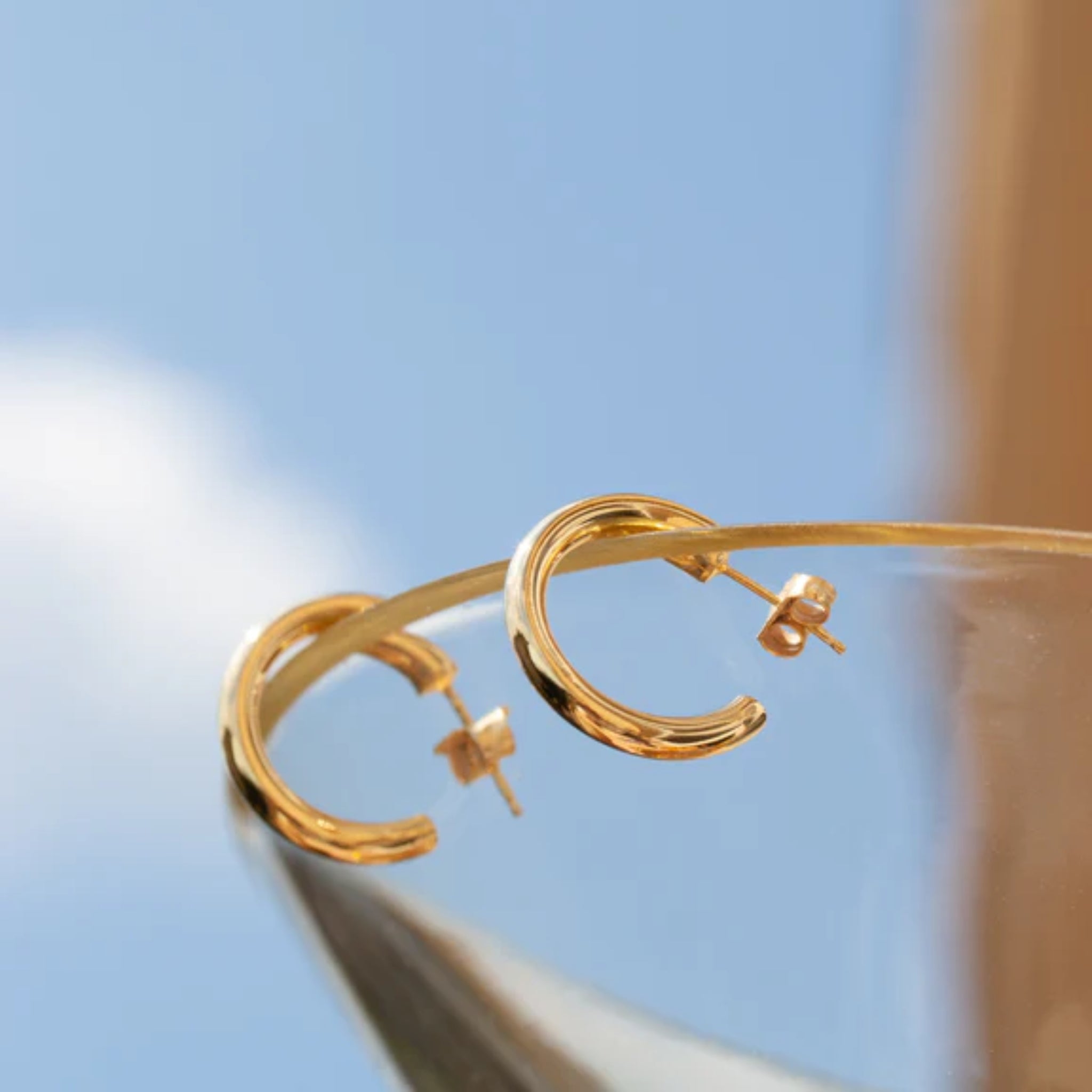 Goldene Creolen auf einem Glas vor blauem Himmel mit weißen Wolken.