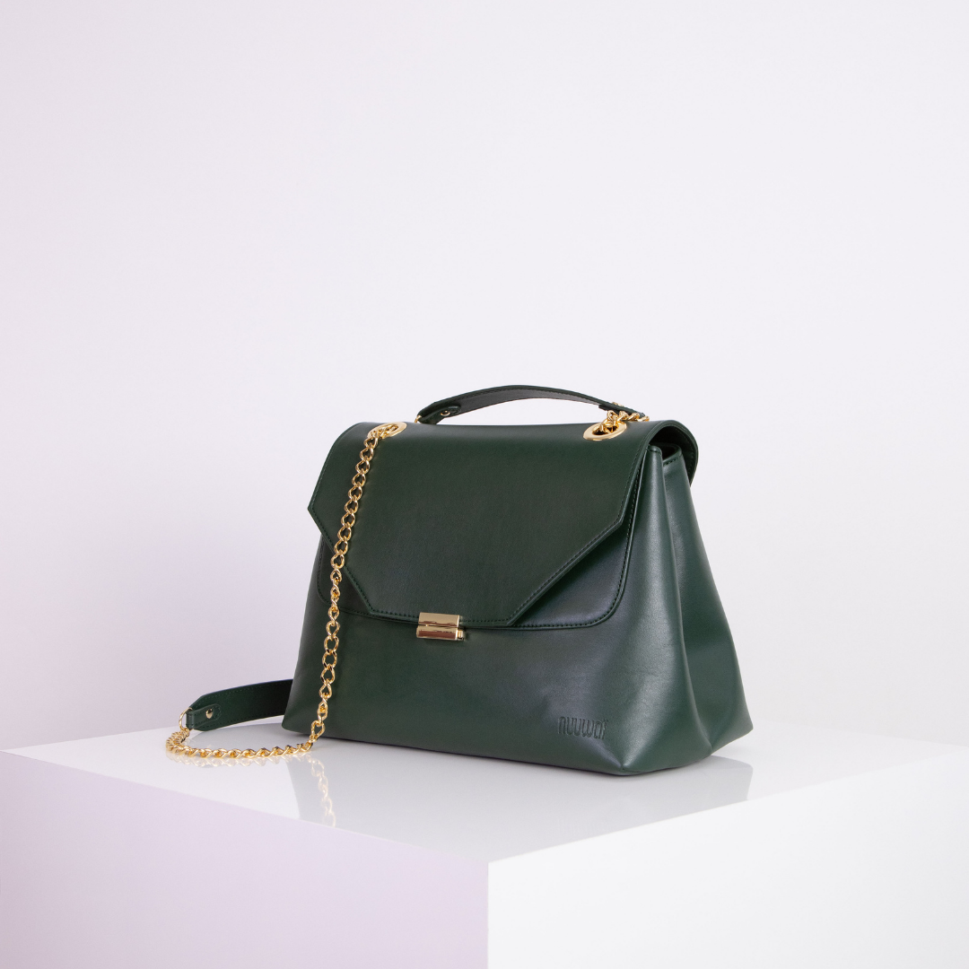 Dunkelgrüne Handtasche mit goldener Kette auf einem weißen Podest vor hellem Hintergrund.