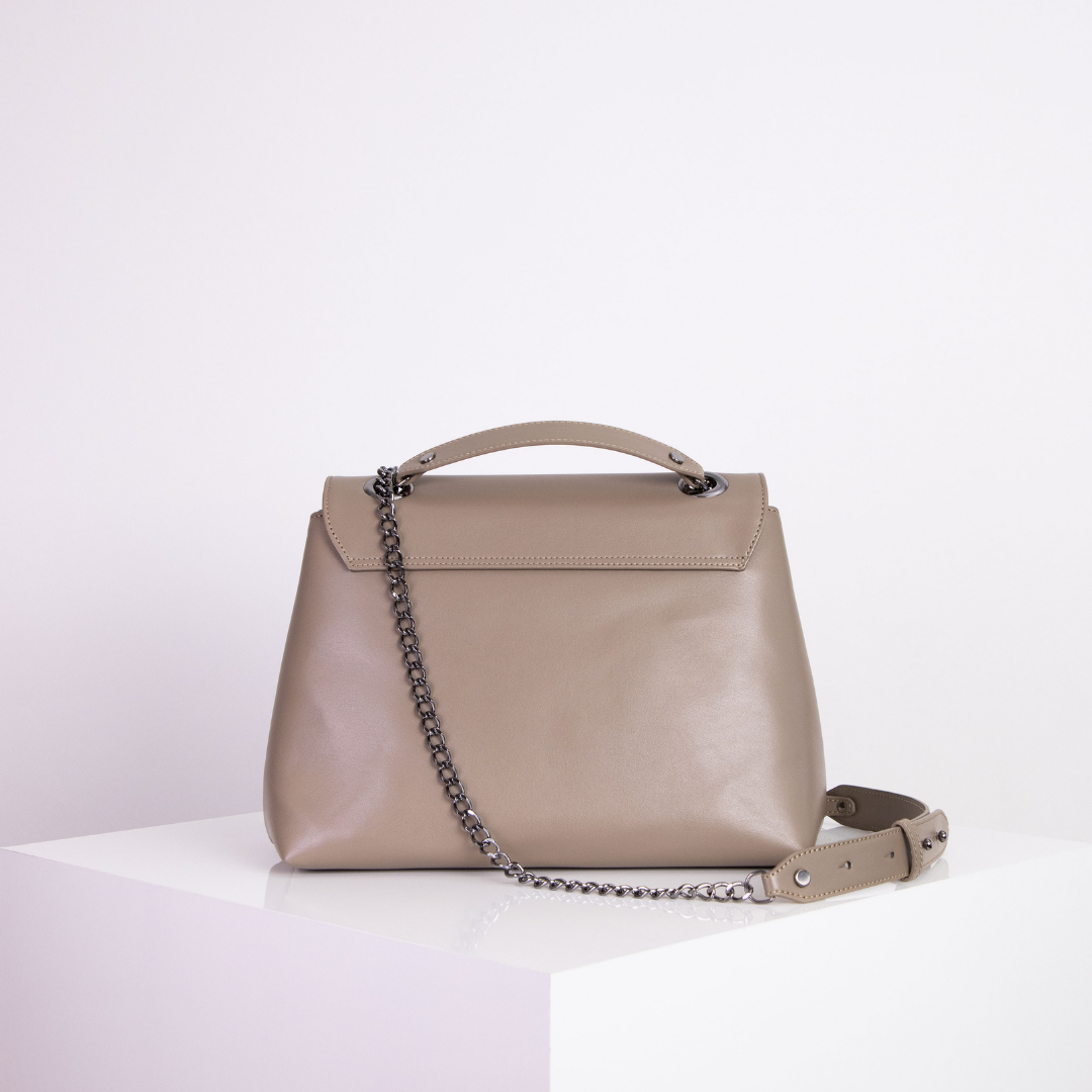 Beige Handtasche mit Kettenriemen auf einem weißen Podest vor neutralem Hintergrund.