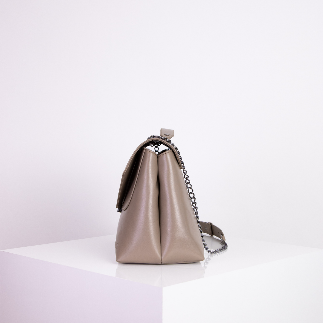 Beige Handtasche aus Leder mit Kettenriemen steht auf einem weißen Podest vor einem hellen Hintergrund.