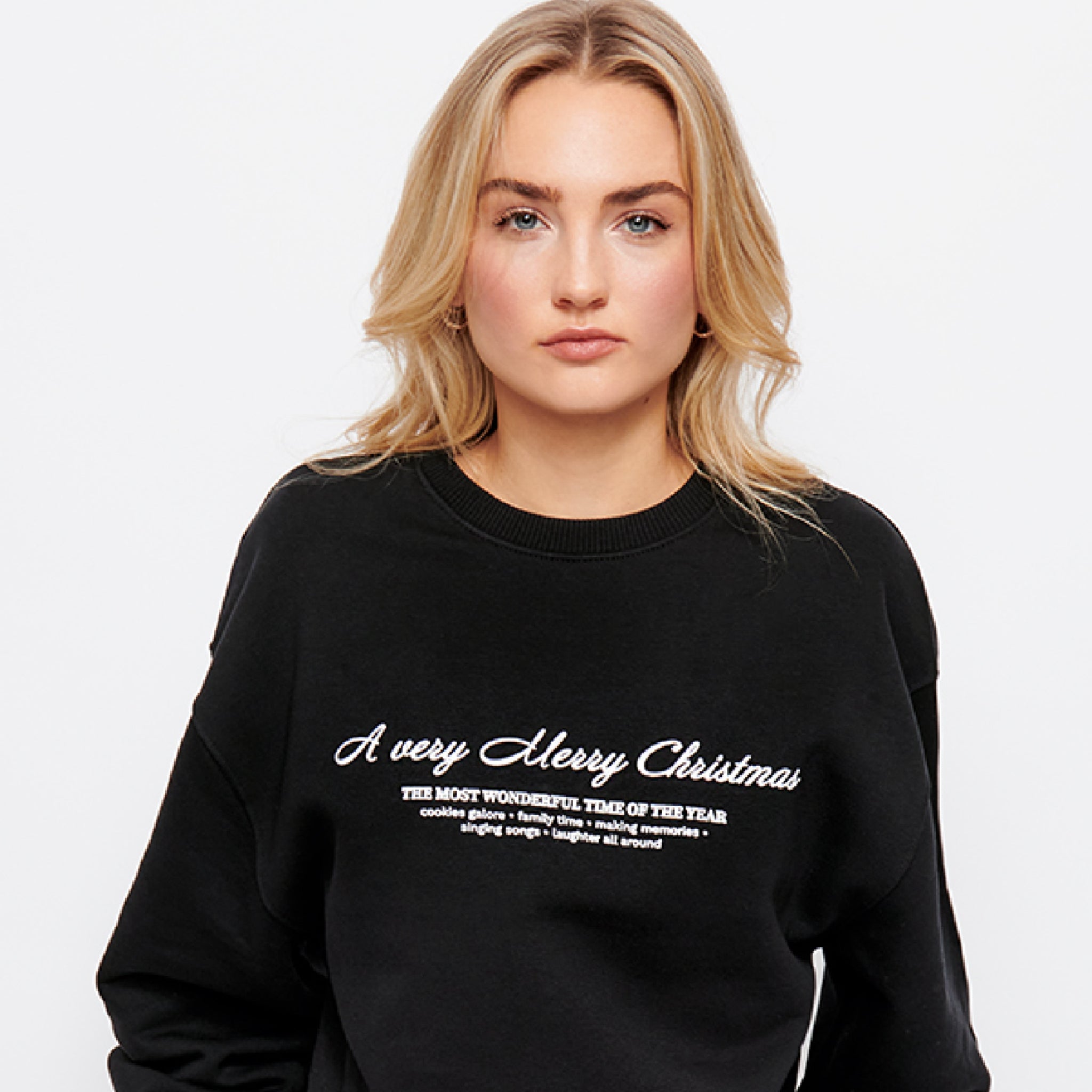 Junge Frau mit blonden Haaren trägt schwarzen Pullover mit der Aufschrift „A very Merry Christmas“ in weißem Text.