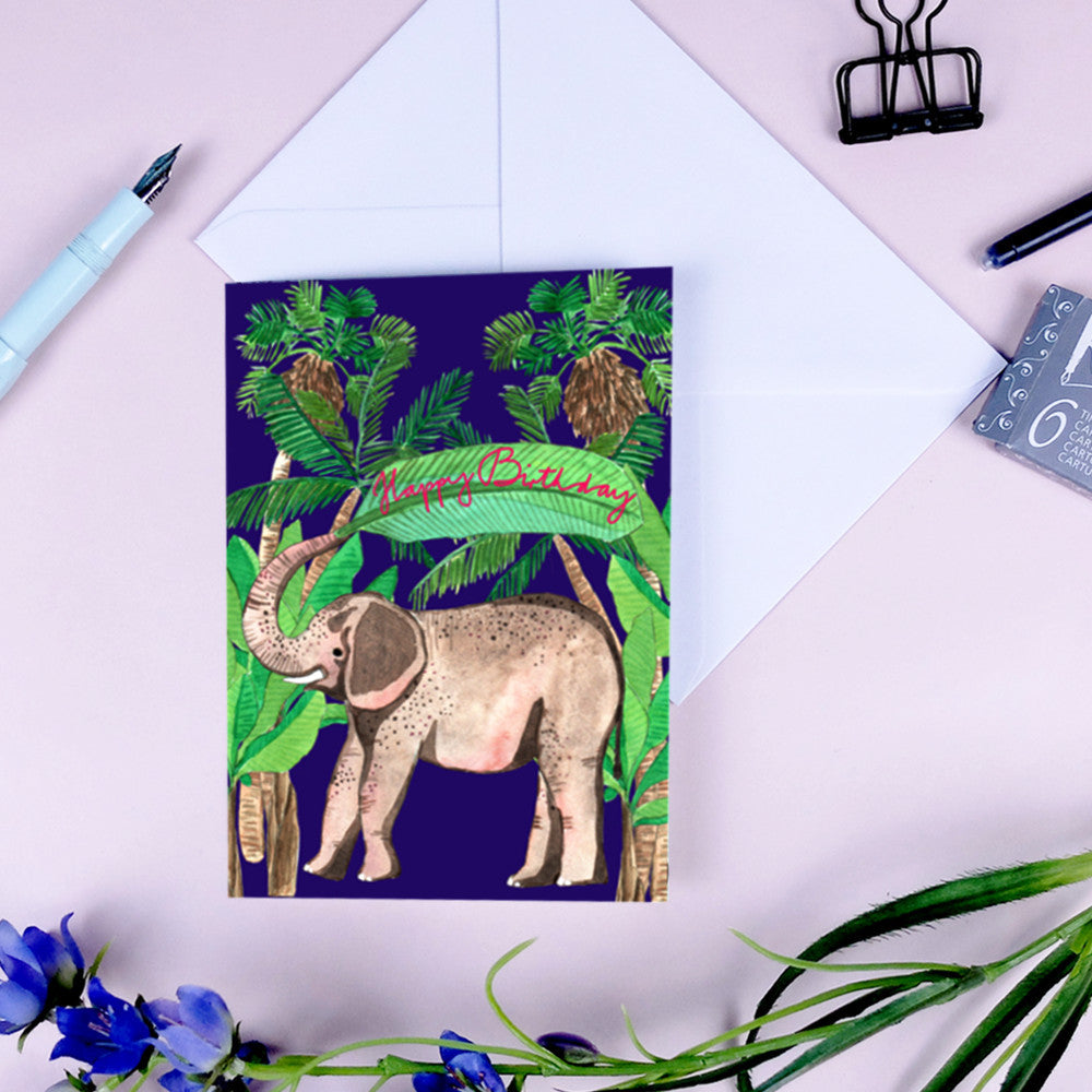 Geburtstagskarte mit Elefant und Palmen, umgeben von Bürobedarf und Blumendekoration auf einem rosa Hintergrund.