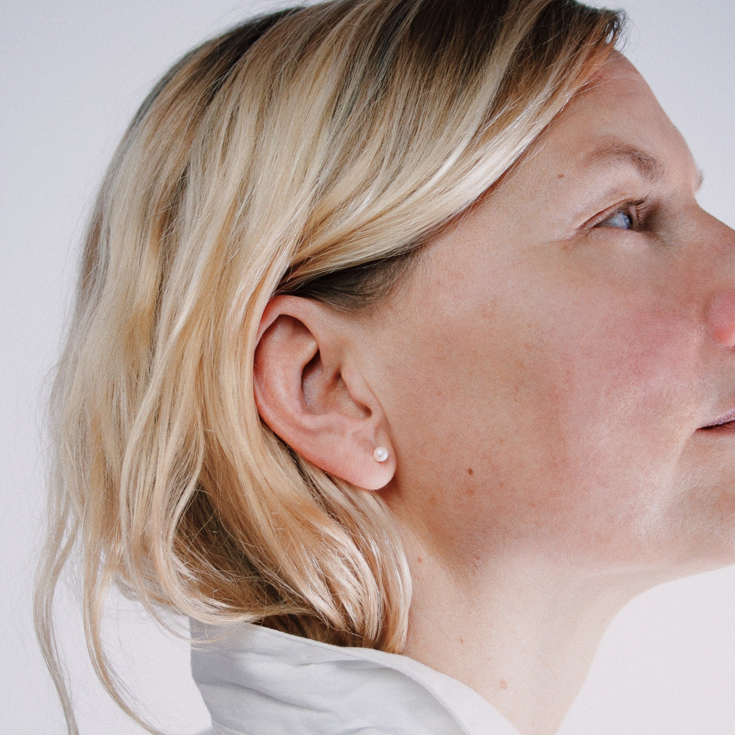 Nahaufnahme einer Frau mit blondem Haar und einem kleinen Perlenohrstecker, die zur Seite schaut.