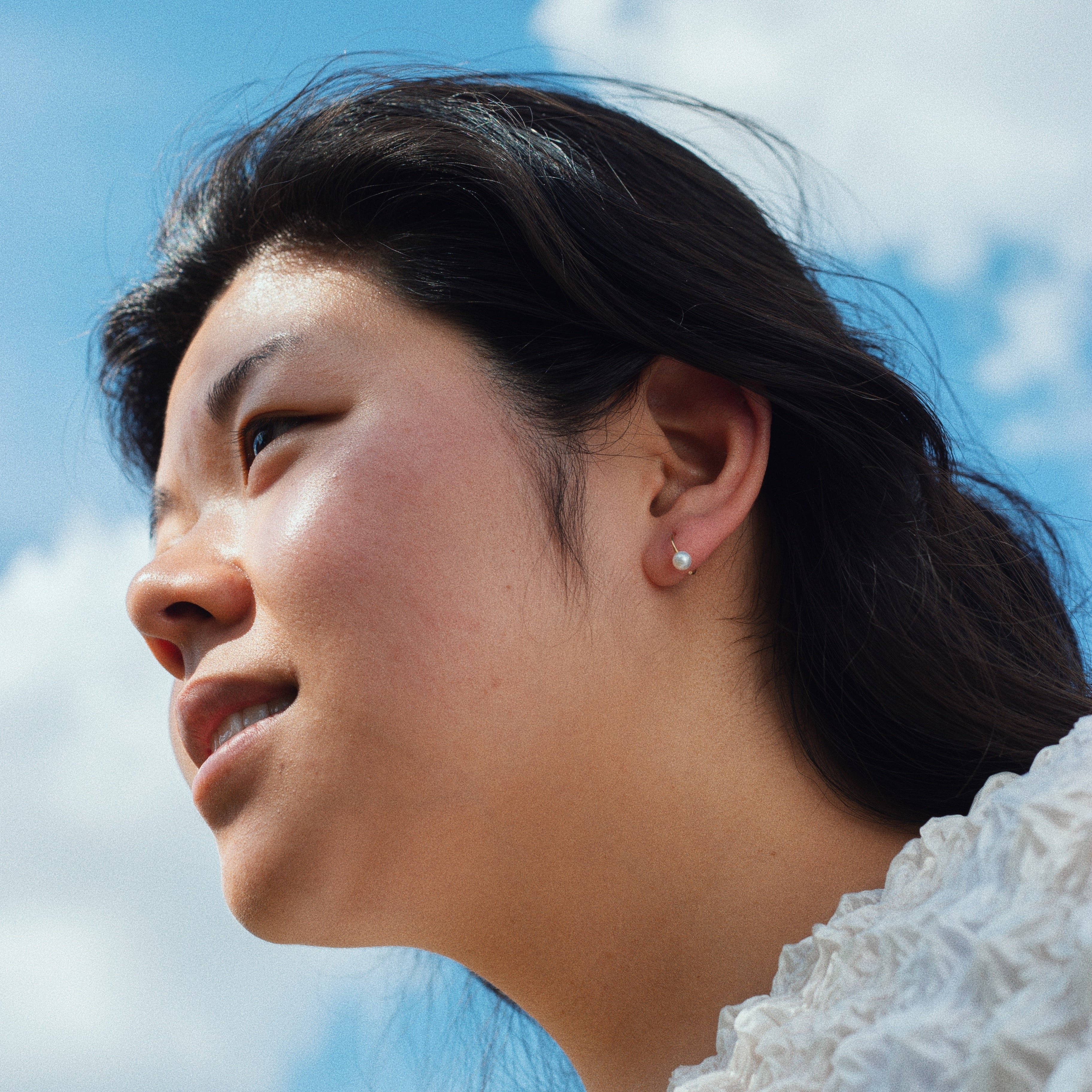 Frau mit langem dunklem Haar blickt nach rechts, vor blauem Himmel mit Wolken. Sie trägt einen weißen Pullover und Ohrringe.