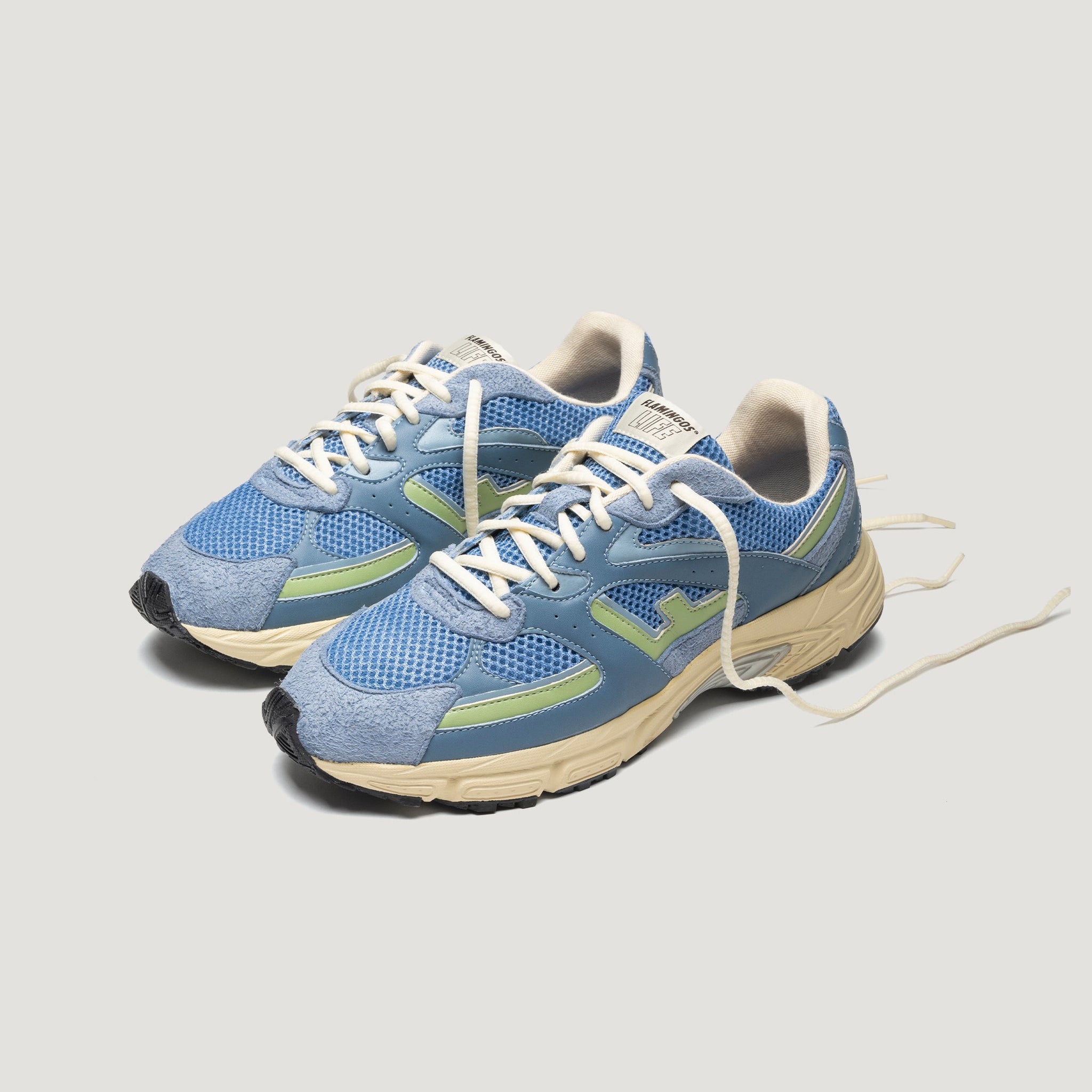 Ein Paar blaue und hellgrüne Sportschuhe mit cremefarbenen Schnürsenkeln und beigen Sohlen, die nebeneinander auf einem schlichten hellen Hintergrund stehen.