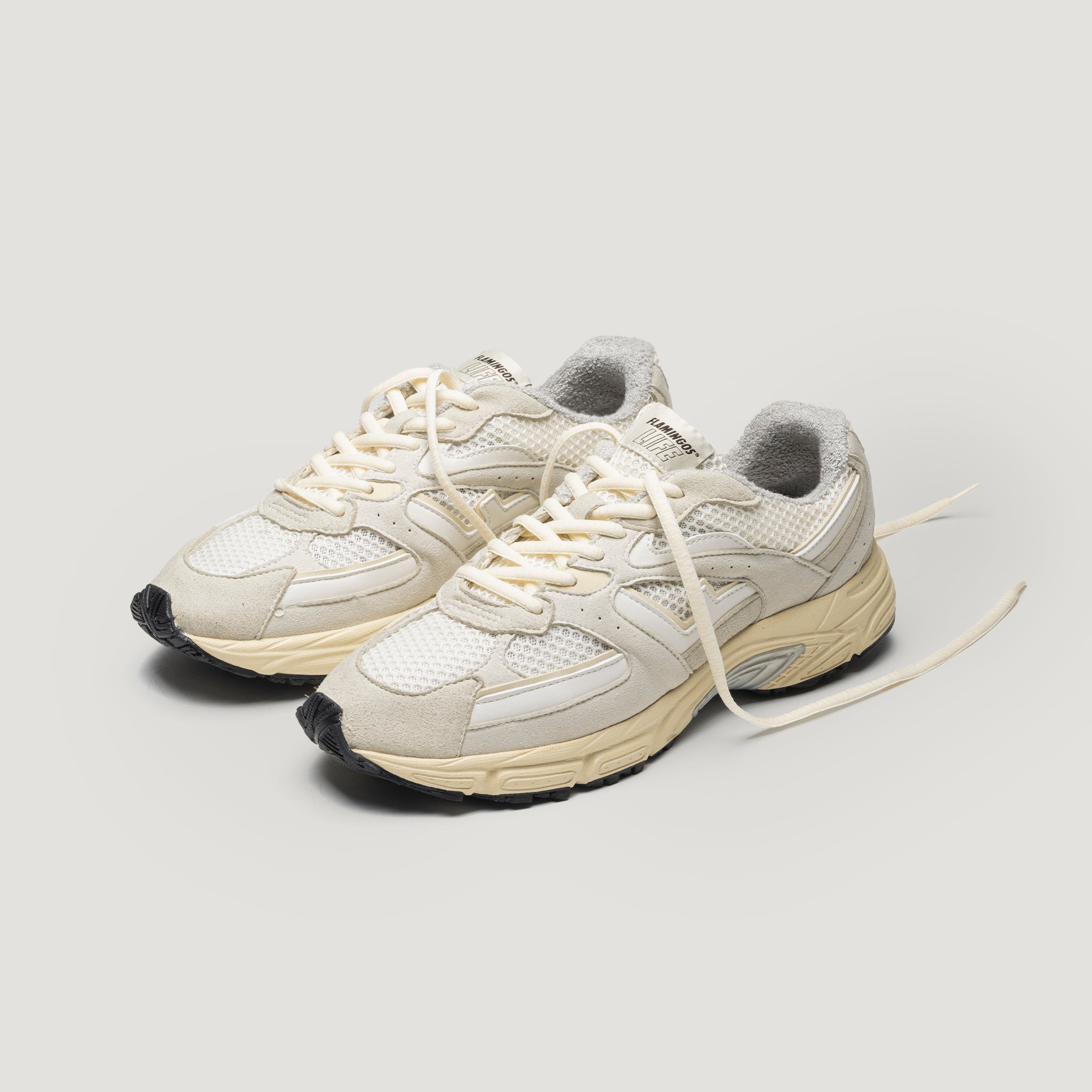 Ein Paar cremefarbene und beigefarbene Sportschuhe mit Mesh-Einsätzen, grauem Innenfutter und weißen Schnürsenkeln auf hellem Hintergrund, wobei die Schnürsenkel nicht gebunden sind.