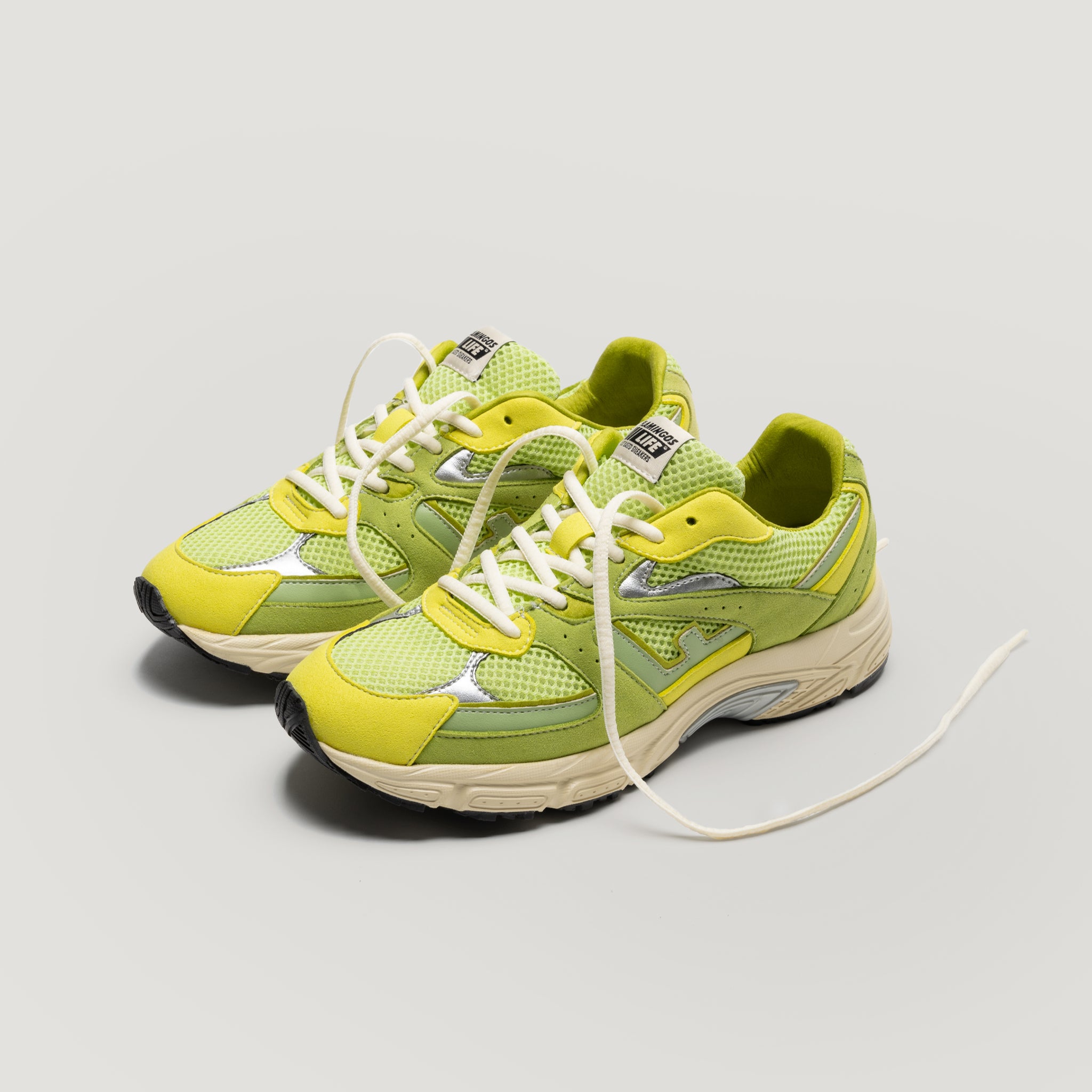 Ein Paar gelbe und grüne Sportschuhe mit weißen Schnürsenkeln, Mesh-Material und klobigen beigen Sohlen ist auf einem schlichten hellen Hintergrund platziert. Ein Schnürsenkel ist locker gelöst und ragt nach außen.