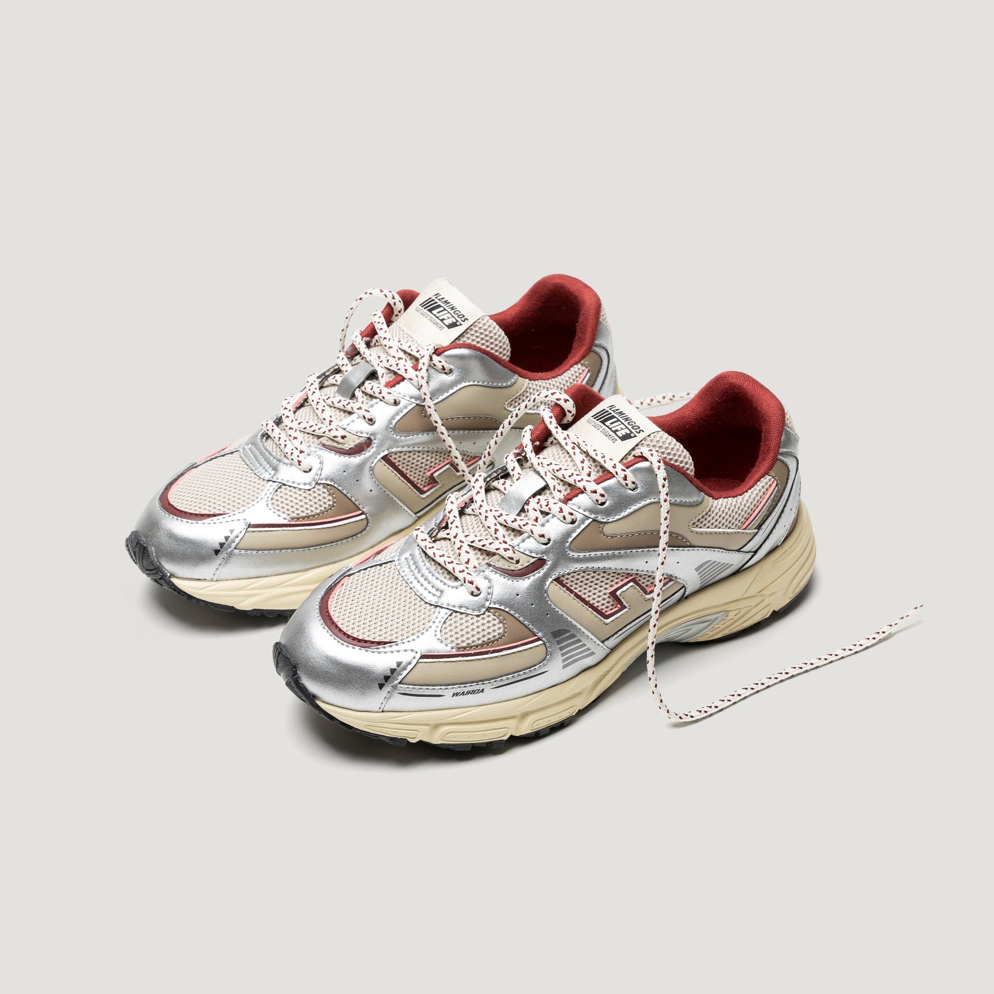 Ein Paar silberne und beige Turnschuhe mit roten Akzenten und weißen Schnürsenkeln, fotografiert auf einem hellgrauen Hintergrund. Ein Schnürsenkel ist gelöst und steht nach außen ab.