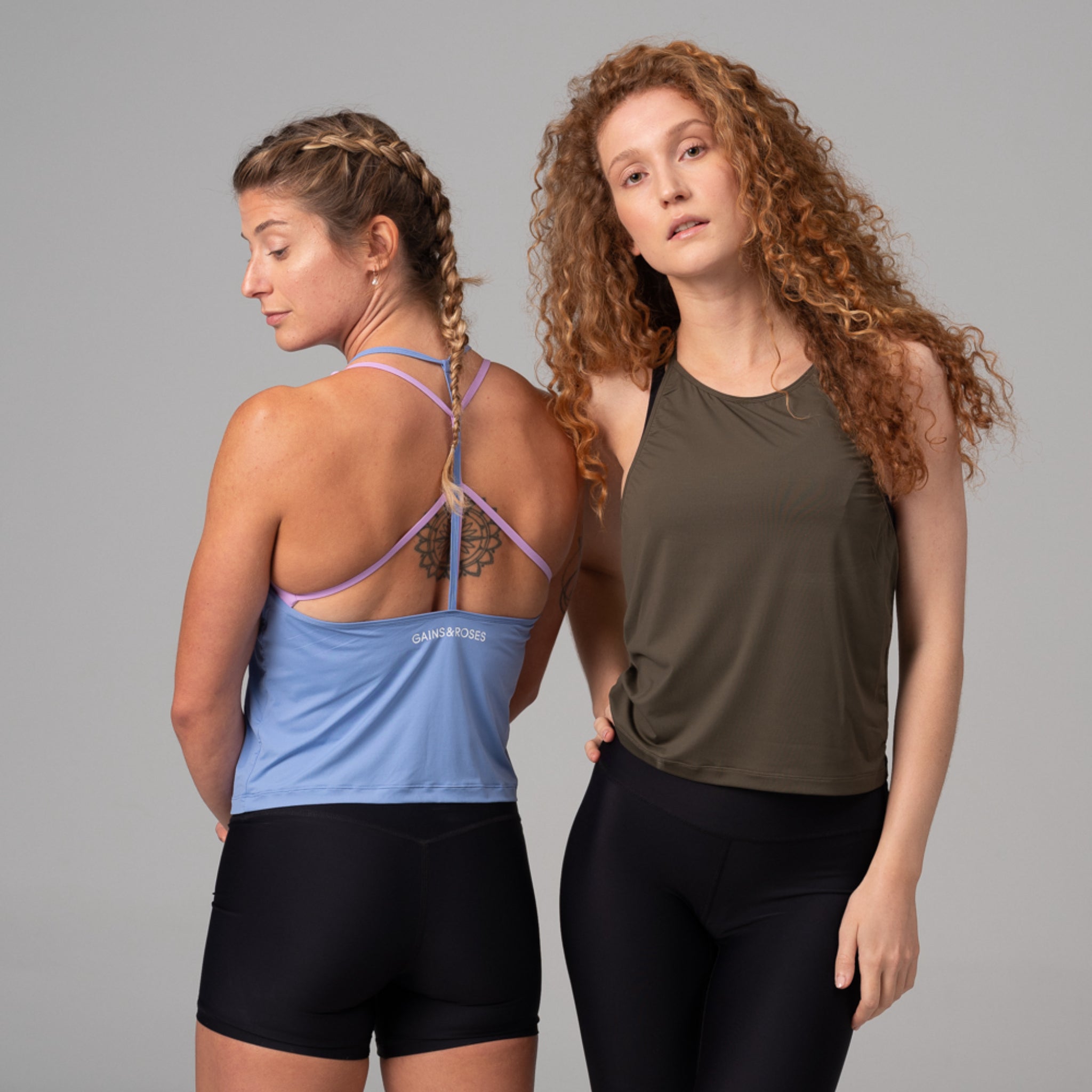 Zwei Frauen in Sportbekleidung, eine mit Zopf und Tattoo, die andere mit lockigem Haar, stehen nebeneinander vor grauem Hintergrund.