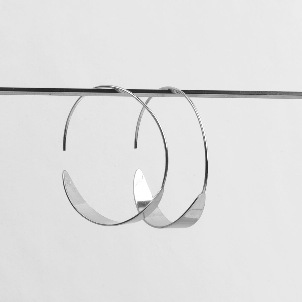 Ein Paar große, minimalistische Silberohrringe mit gedrehtem Muster, die an einem dünnen horizontalen Stab vor einem schlichten weißen Hintergrund hängen.