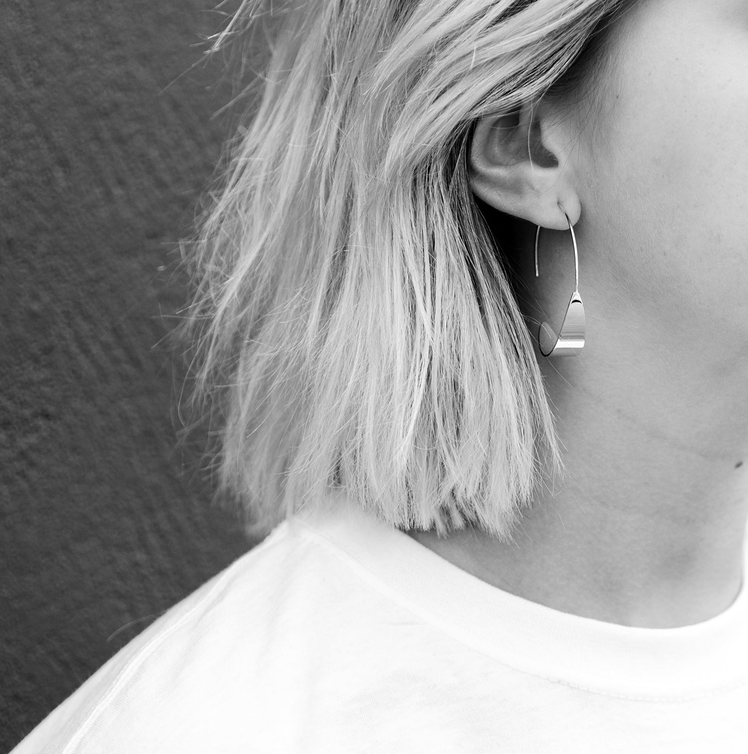 Schwarz-Weiß-Foto einer Person mit kurzen, hellen Haaren, die einen geometrischen Ohrring und ein schlichtes weißes Hemd trägt, dargestellt im Profil vor einem strukturierten dunklen Hintergrund.