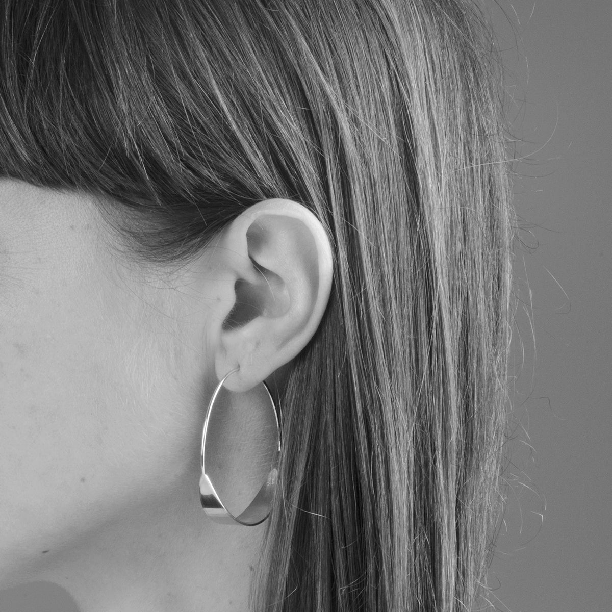 Schwarz-Weiß-Nahaufnahme des Kopfes einer Frau im Profil, die ihr glattes Haar, ihr Ohr und einen großen, runden Reifohrring zeigt. Der Hintergrund ist schlicht und unscharf.