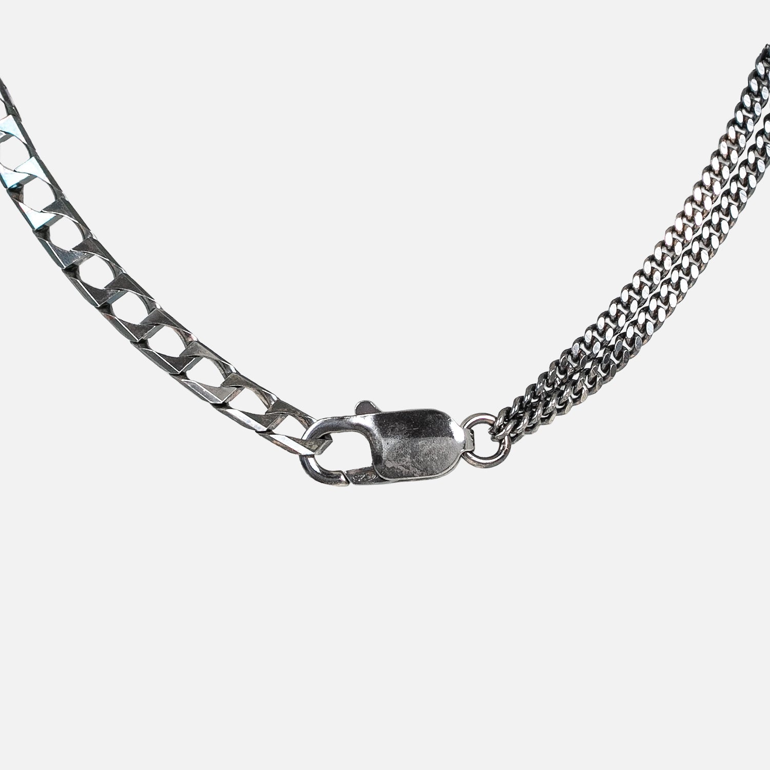 Nahaufnahme einer silbernen Halskette mit Karabinerverschluss, die zwei verschiedene Kettentypen aufweist: eine flache, rechteckige Gliederkette auf der linken Seite und eine feinere, mehrsträngige Kette auf der rechten Seite, vor einem weißen Hintergrund.