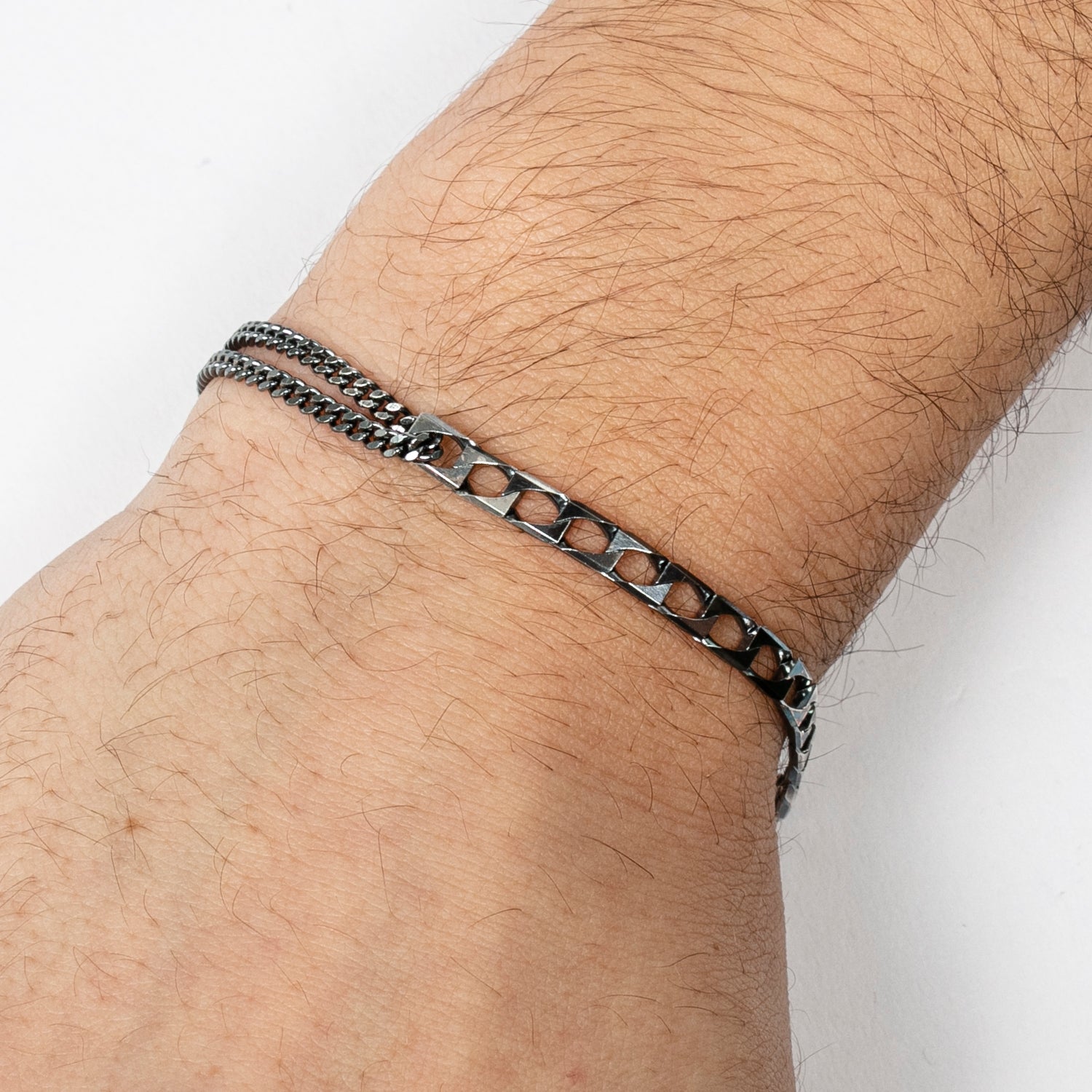 Nahaufnahme eines Handgelenks, das ein dünnes, silbernes Kettenarmband mit einem geometrischen, ausgeschnittenen Muster an einem hellhäutigen Arm trägt, vor einem weißen Hintergrund.