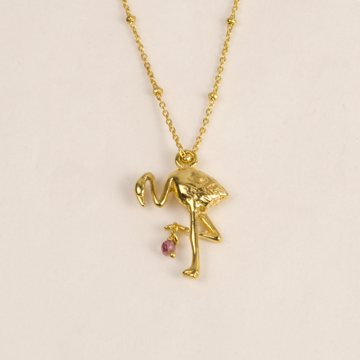 Eine goldene Halskette mit einem Anhänger in Form eines Flamingos und einem kleinen rosafarbenen Edelstein, der unter einem Bein hängt, vor einem schlichten, hellen Hintergrund.