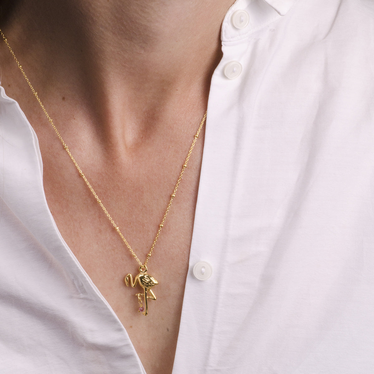 Eine Person, die ein weißes Hemd mit aufgeknöpften Knöpfen trägt, an dem eine goldene Halskette mit einem Flamingo und dem Buchstaben N hängt.