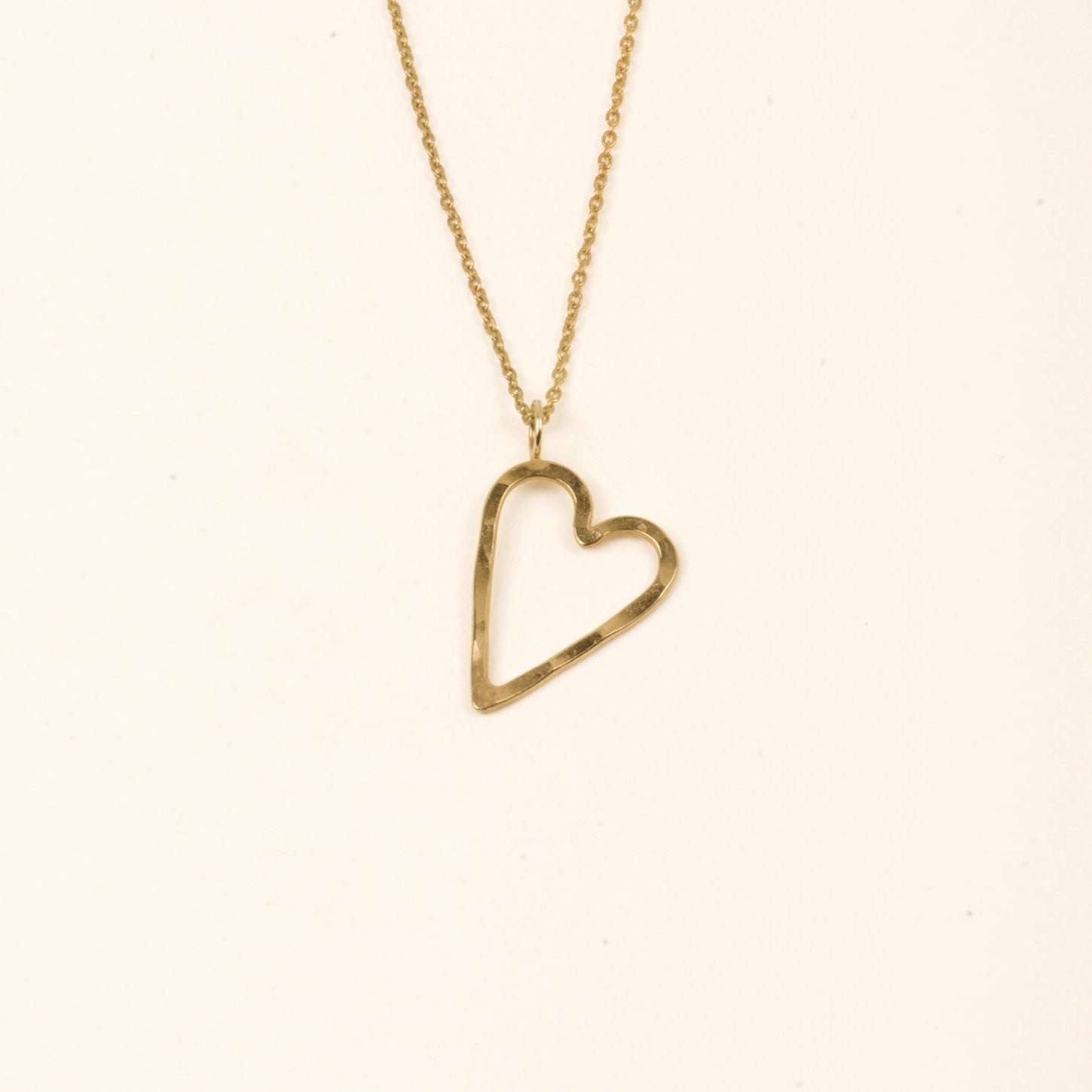 Eine goldene Halskette mit einer zarten Kette und einem offenen, asymmetrischen herzförmigen Anhänger hängt vor einem schlichten, hellen Hintergrund.