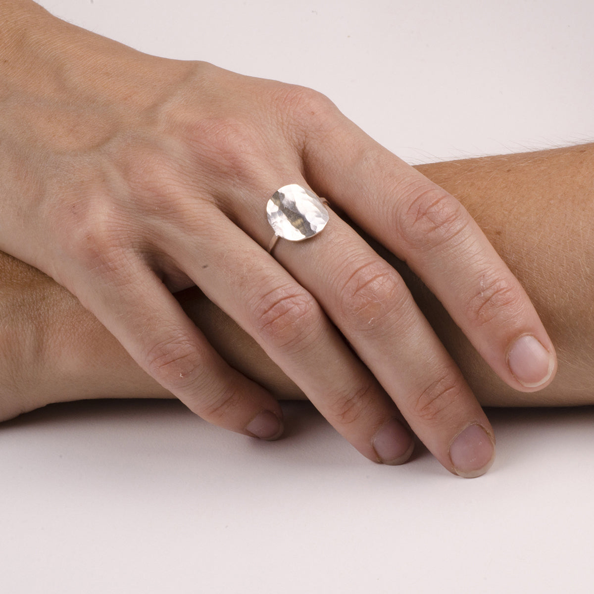 Eine Hand mit Naturnägeln ruht auf einem Arm und trägt einen großen, runden, gehämmerten Silberring am Mittelfinger. Der Hintergrund ist einfarbig weiß.