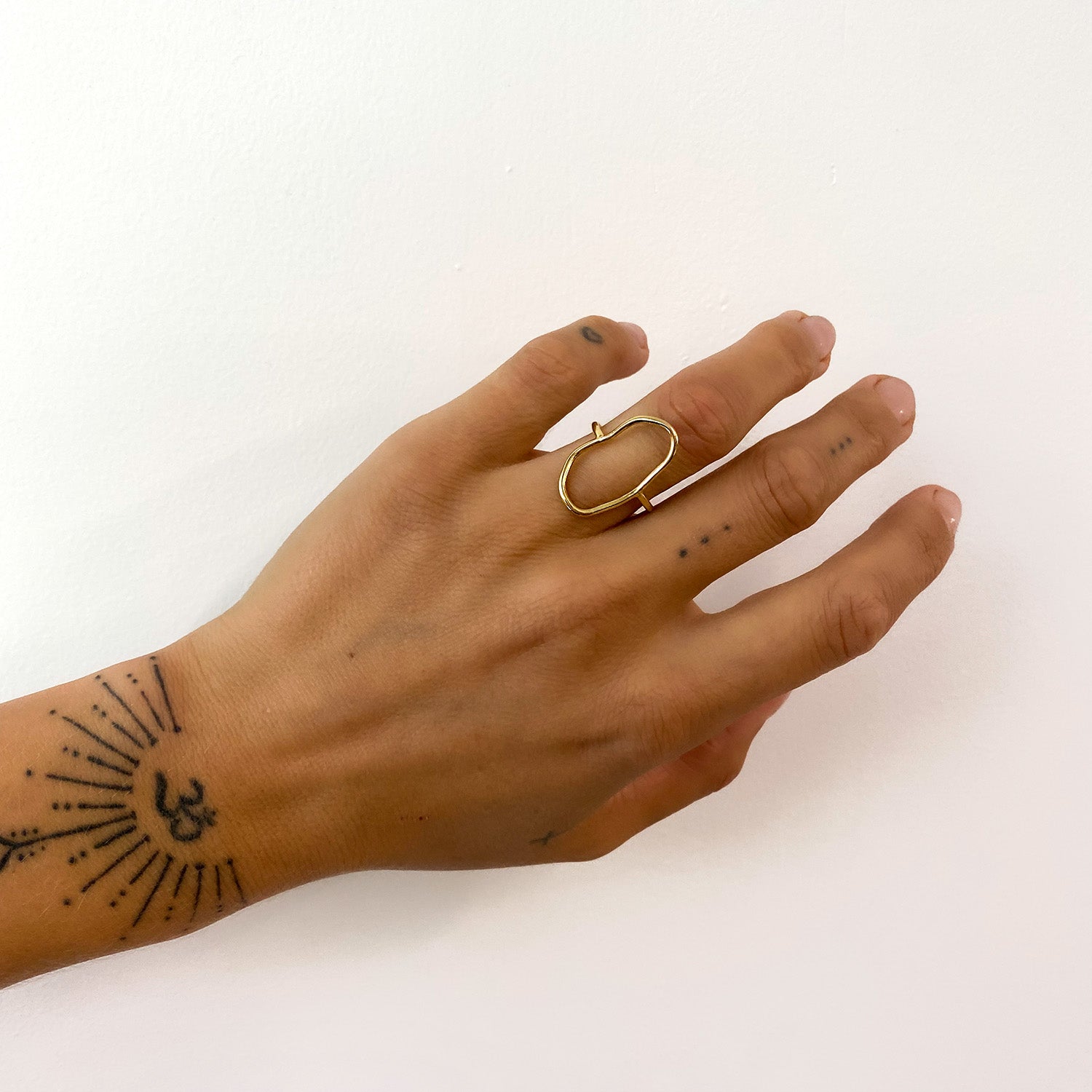 Eine Hand mit einer Tätowierung mit Sonnenschlange und Om-Symbol trägt einen großen, unregelmäßig geformten Goldring am Mittelfinger. An den Fingern befinden sich außerdem kleine Punkttattoos. Der Hintergrund ist eine schlichte weiße Wand.