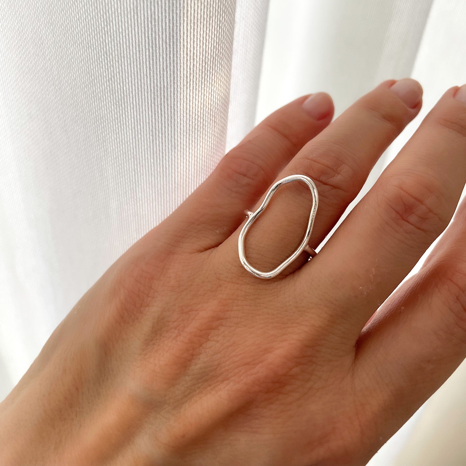 Eine Hand, die einen minimalistischen, unregelmäßig geformten Silberring trägt, wird vor einem sanft beleuchteten weißen Vorhang gezeigt.