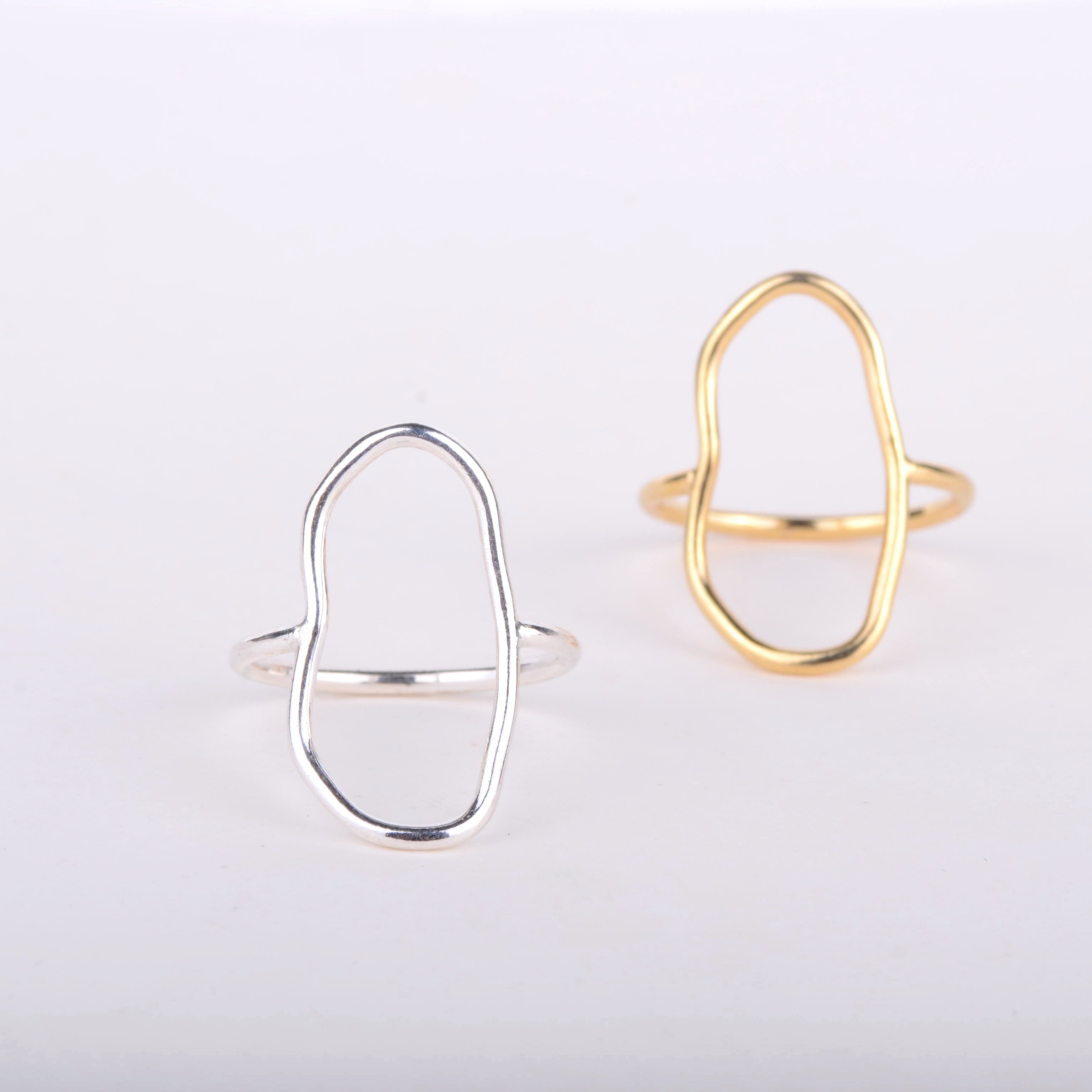 Zwei minimalistische Ringe sind auf einem weißen Hintergrund abgebildet. Ein Ring ist aus Silber und der andere aus Gold. Beide haben ein offenes, asymmetrisches, ovales Design als Herzstück.