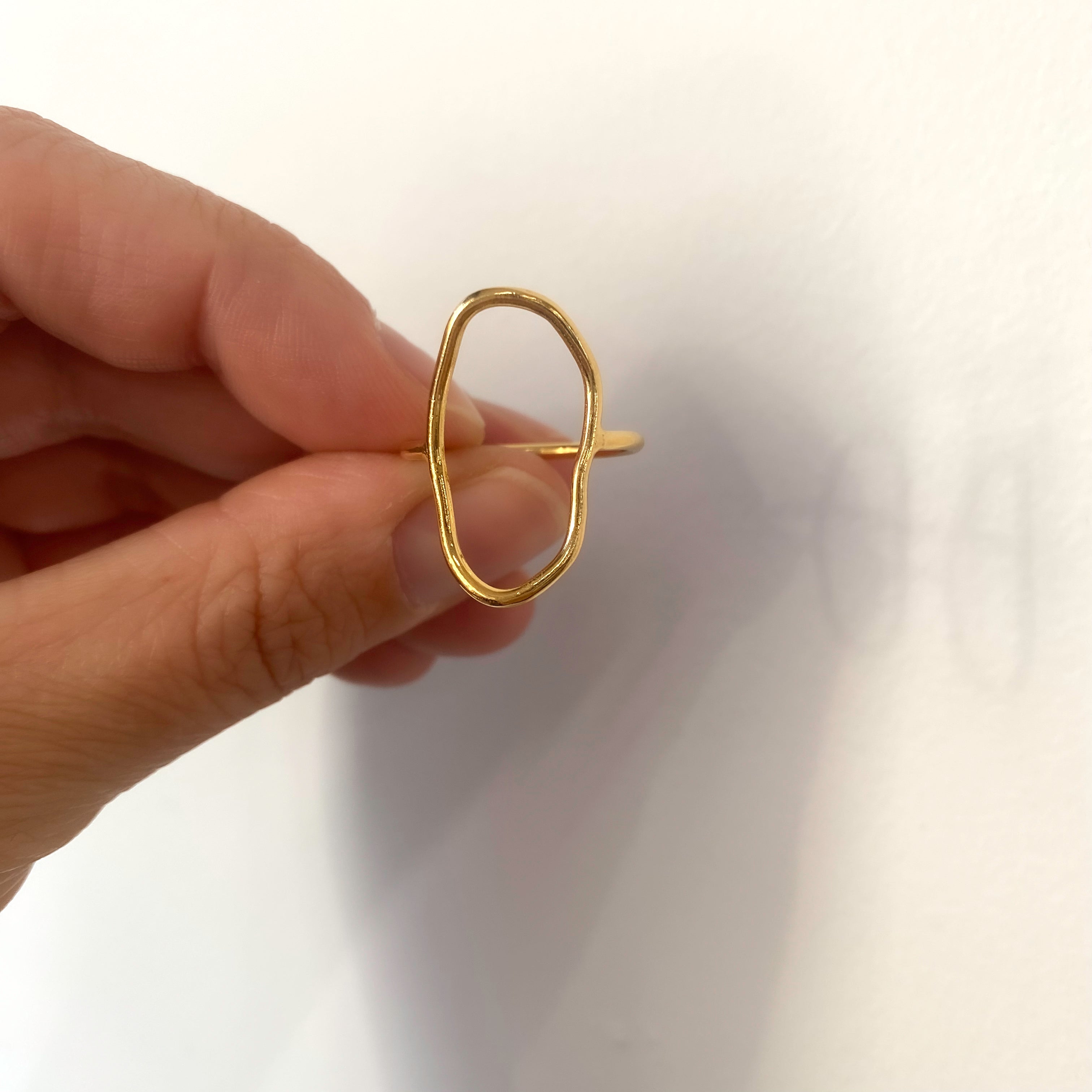 Eine Hand hält einen minimalistischen Goldring mit einer unregelmäßigen, organisch geformten, offenen Kontur vor einem schlichten weißen Hintergrund.