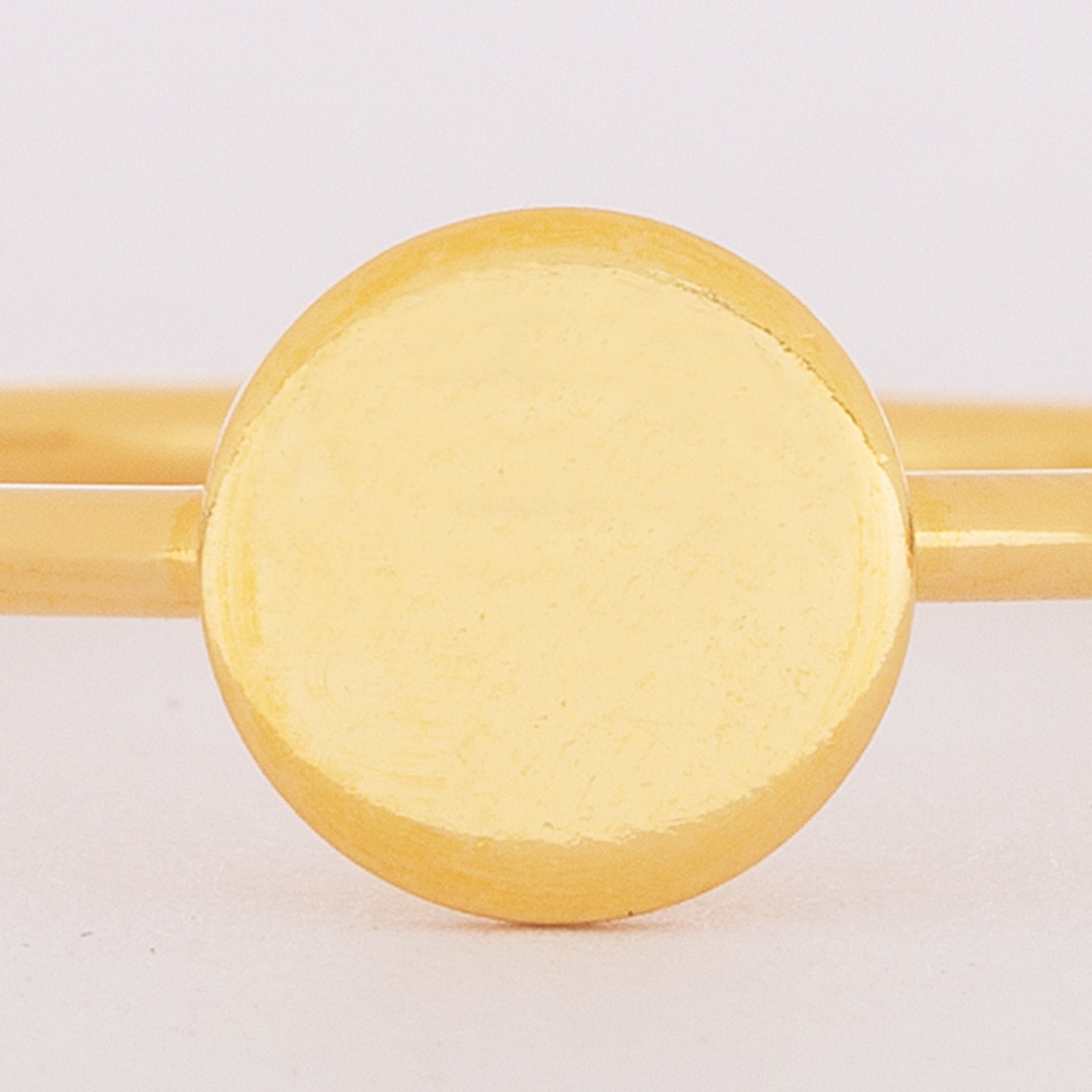 Nahaufnahme eines goldenen Armreifs mit einem glatten, flachen, kreisförmigen Mittelstück mit glänzender Oberfläche vor einem schlichten, hellen Hintergrund.