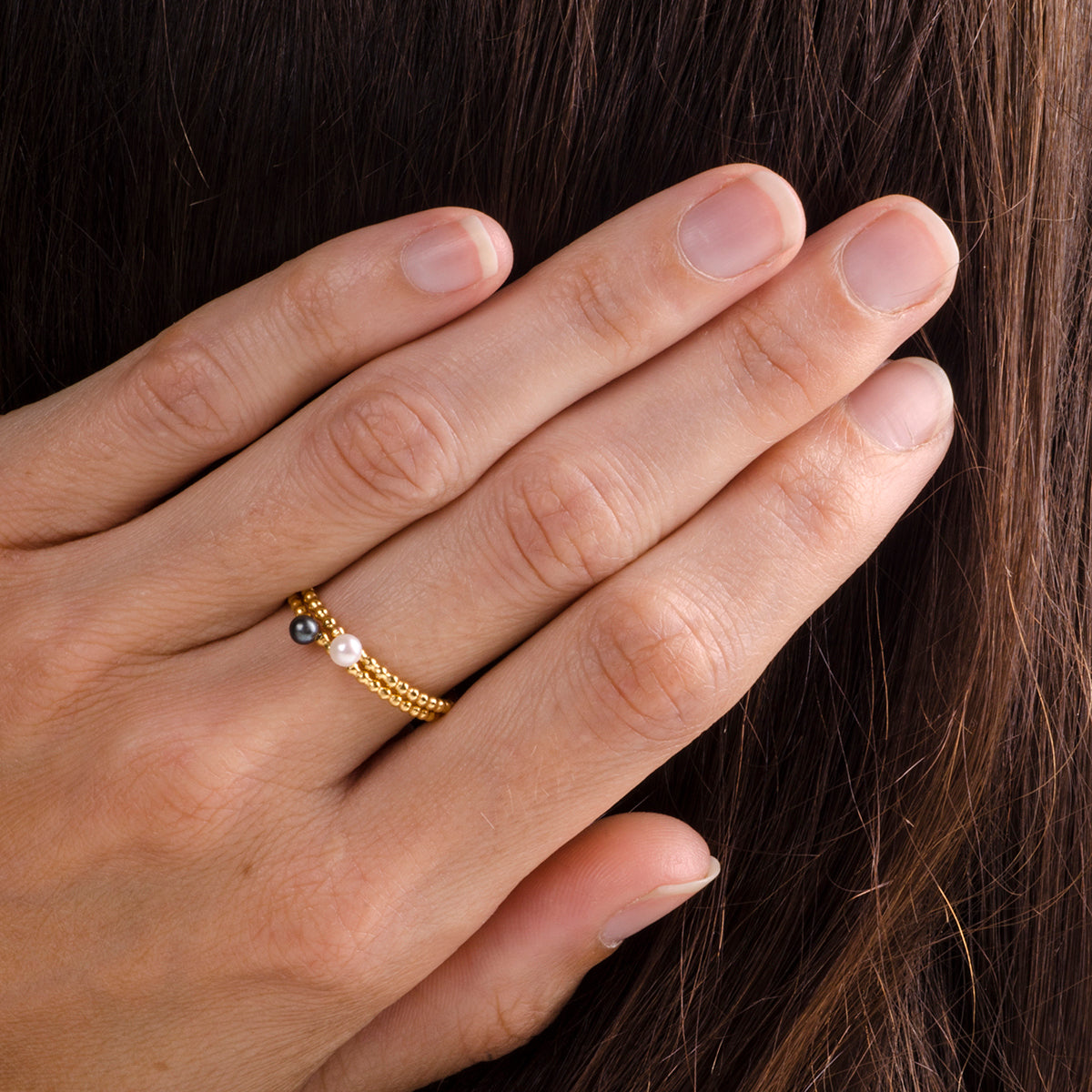 Nahaufnahme einer Hand mit sauber geschnittenen Nägeln, die auf langem braunem Haar ruht. Sie trägt einen zarten Goldring, der mit drei kleinen Perlen in Schwarz, Weiß und Gold verziert ist.