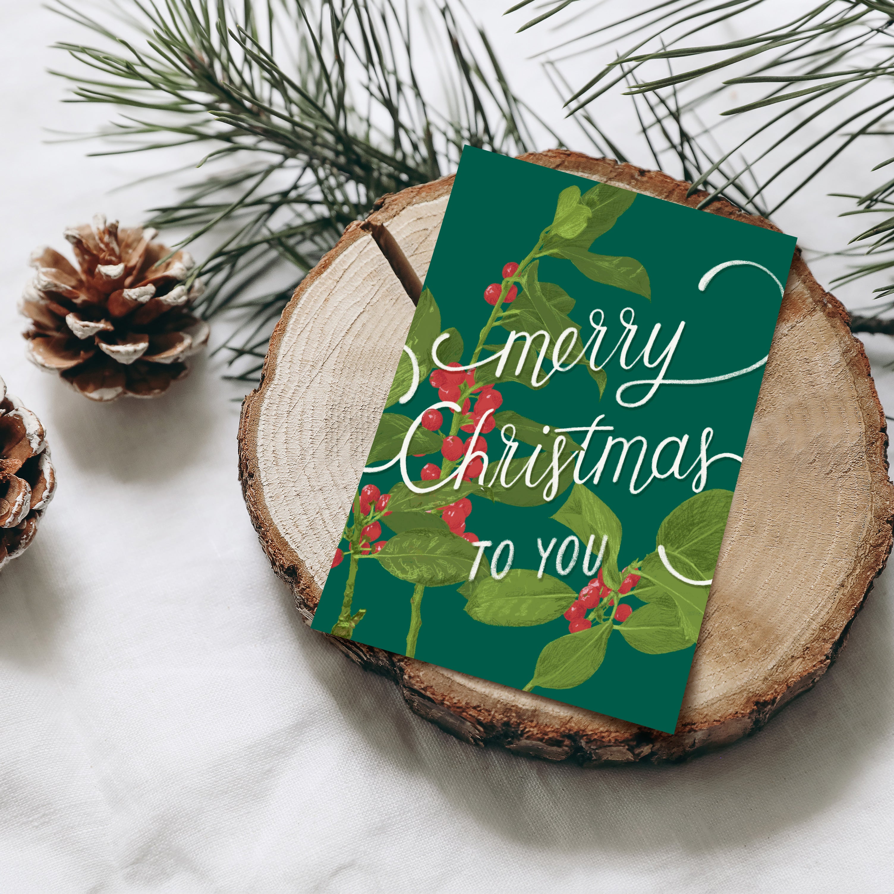 Eine festliche "Merry Christmas to You"-Karte mit Stechpalmen und Beeren liegt auf einer Holzscheibe, umgeben von Tannenzweigen und einem Tannenzapfen auf einem weißen Stoffhintergrund.