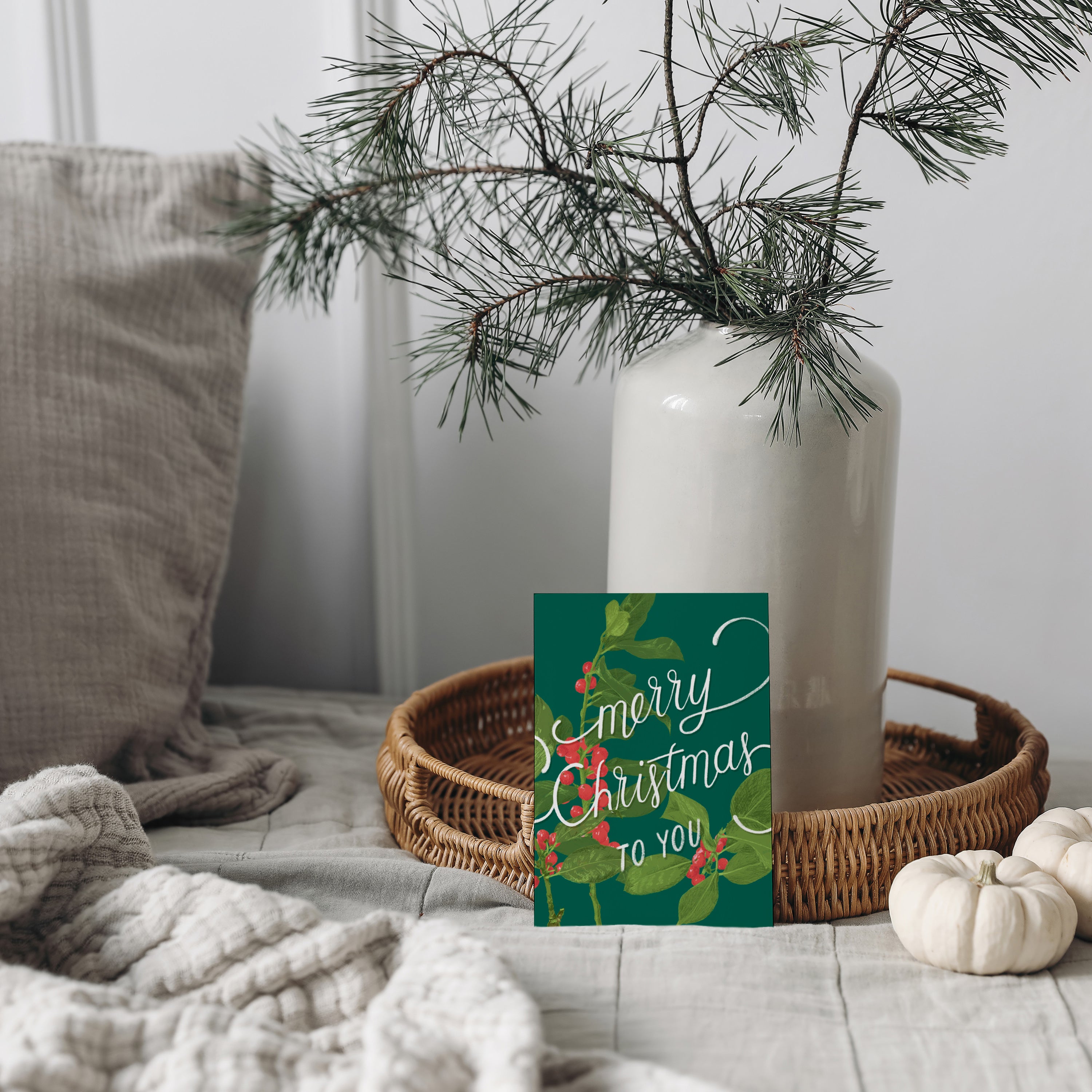 Ein Korbtablett mit einer weißen Vase mit Tannenzweigen und einer Weihnachtskarte mit der Aufschrift "Frohe Weihnachten" steht auf einem gemütlichen Bett mit einer beigen Strickdecke und weichen Kissen.