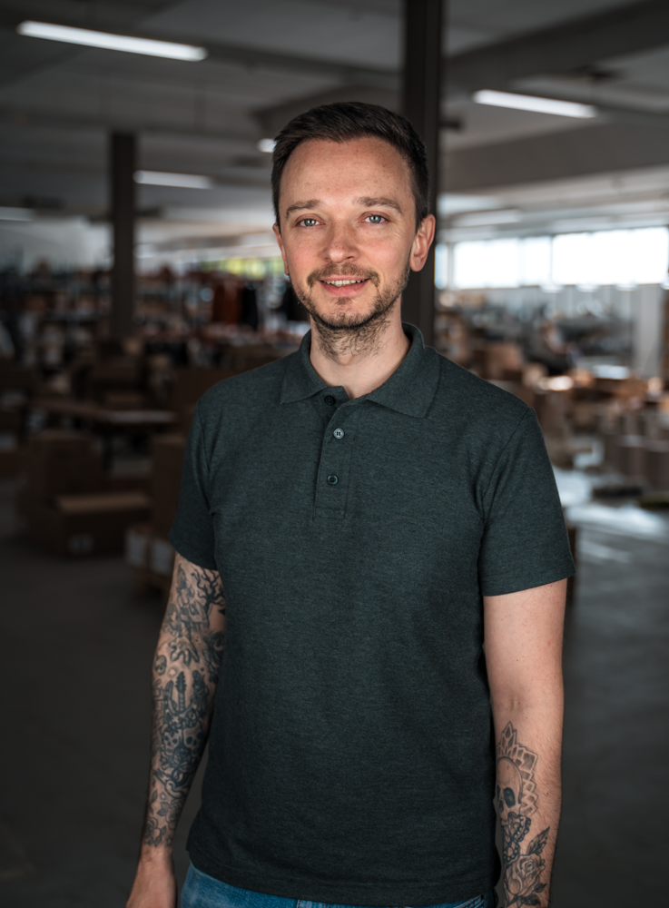 Ein Mann mit Tätowierungen steht lächelnd in einem Lagerraum.