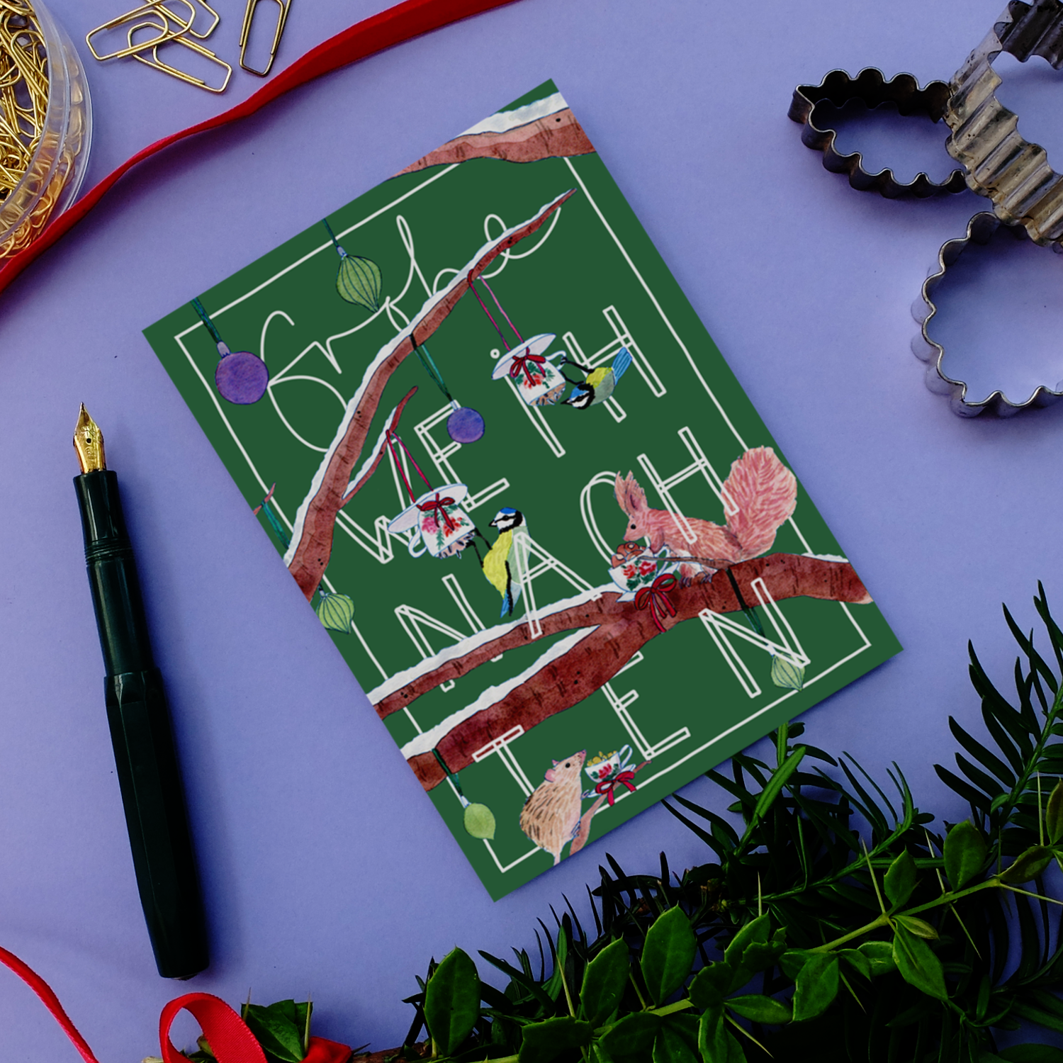 Eine festliche Karte mit Frohe Weihnachten in weißer Schrift auf grünem Grund, verziert mit Vögeln, Geschenken und Ornamenten auf Baumzweigen. Umgeben von einem Stift, Ausstechformen, goldenen Büroklammern und Grünzeug auf einer lila Fläche.