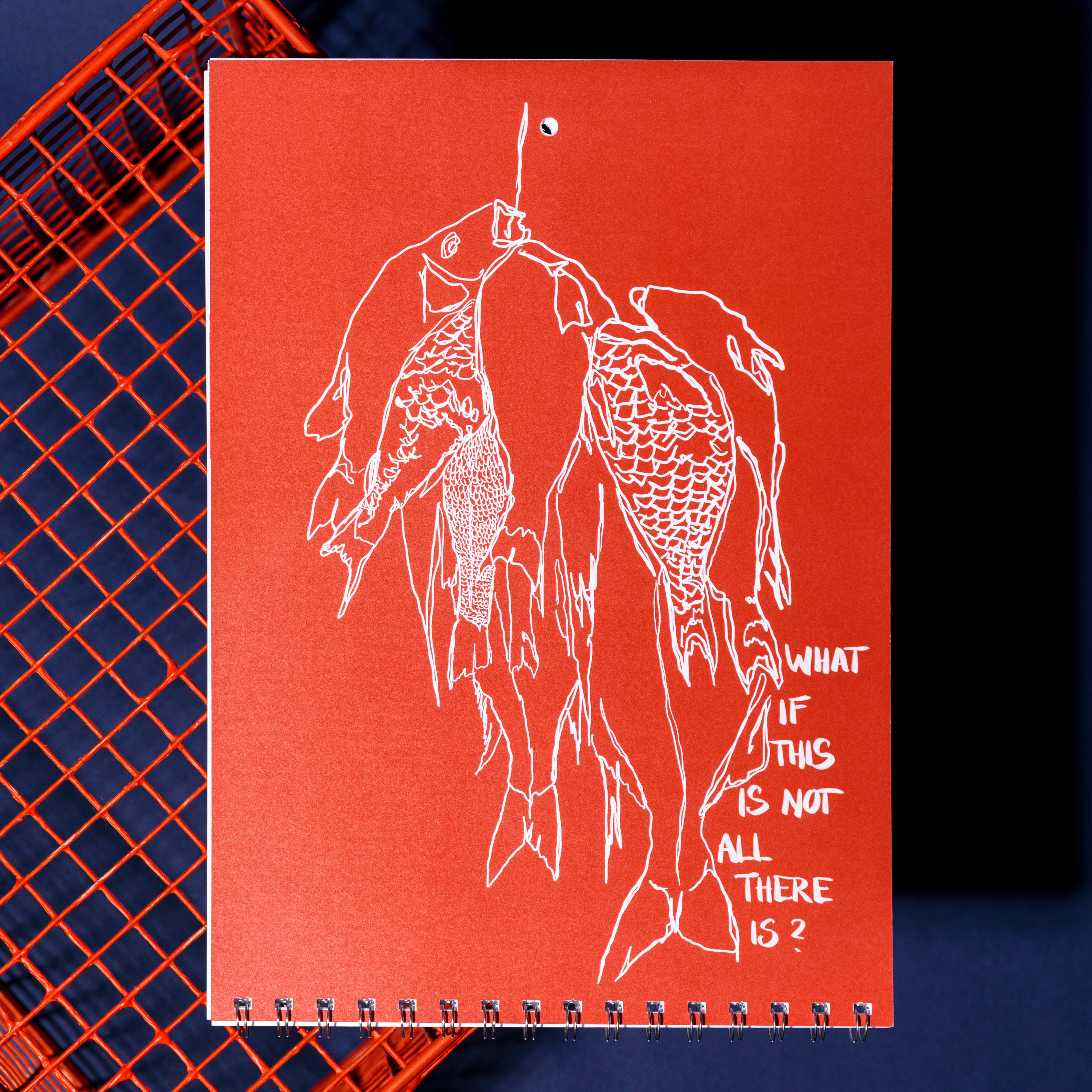 Ein rotes, spiralgebundenes Notizbuch mit einer weißen Strichzeichnung von mehreren hängenden Fischen und dem handgeschriebenen Text WHAT IF THIS IS NOT ALL THERE IS? auf dem Einband. Das Notizbuch liegt auf einer roten Drahtgitterfläche.