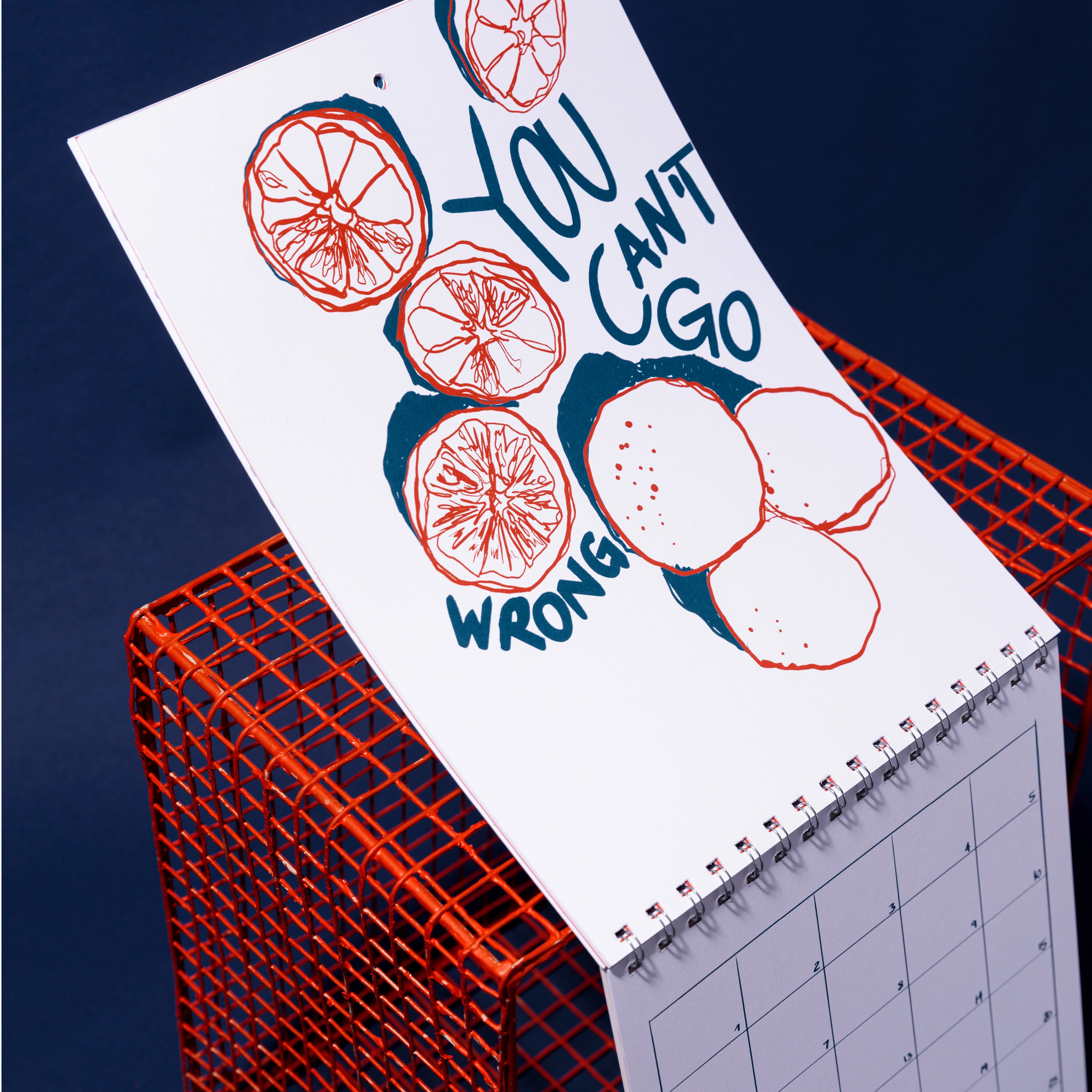 Ein spiralgebundener Kalender mit abstrakten orangefarbenen und blauen Illustrationen und dem Schriftzug YOU CANT GO WRONG befindet sich auf einem roten Drahtgitterwürfel vor einem dunkelblauen Hintergrund.