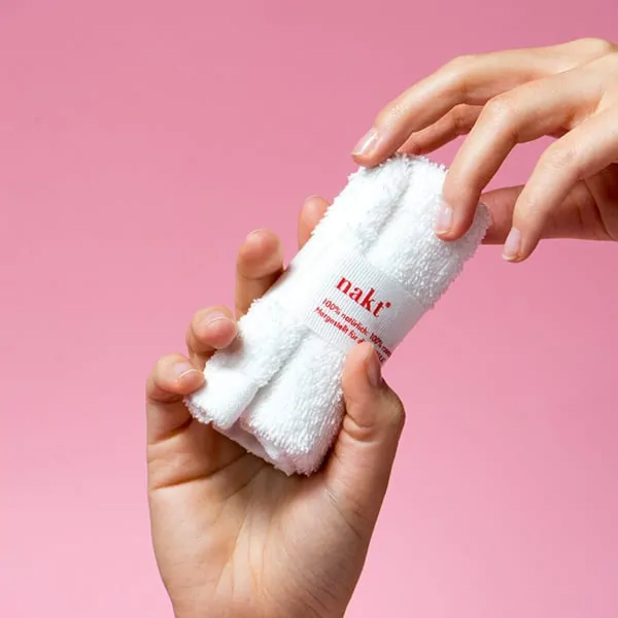 Hände halten ein zusammengerolltes weißes Handtuch mit dem Aufdruck "nakt." vor einem rosa Hintergrund.