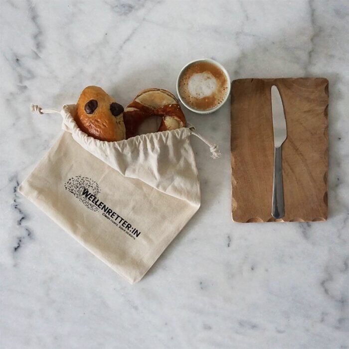Eine Stofftasche mit Brötchen und Brezeln liegt auf einer Marmorplatte neben einem Holzbrett mit einem Buttermesser und einer Tasse Kaffee.